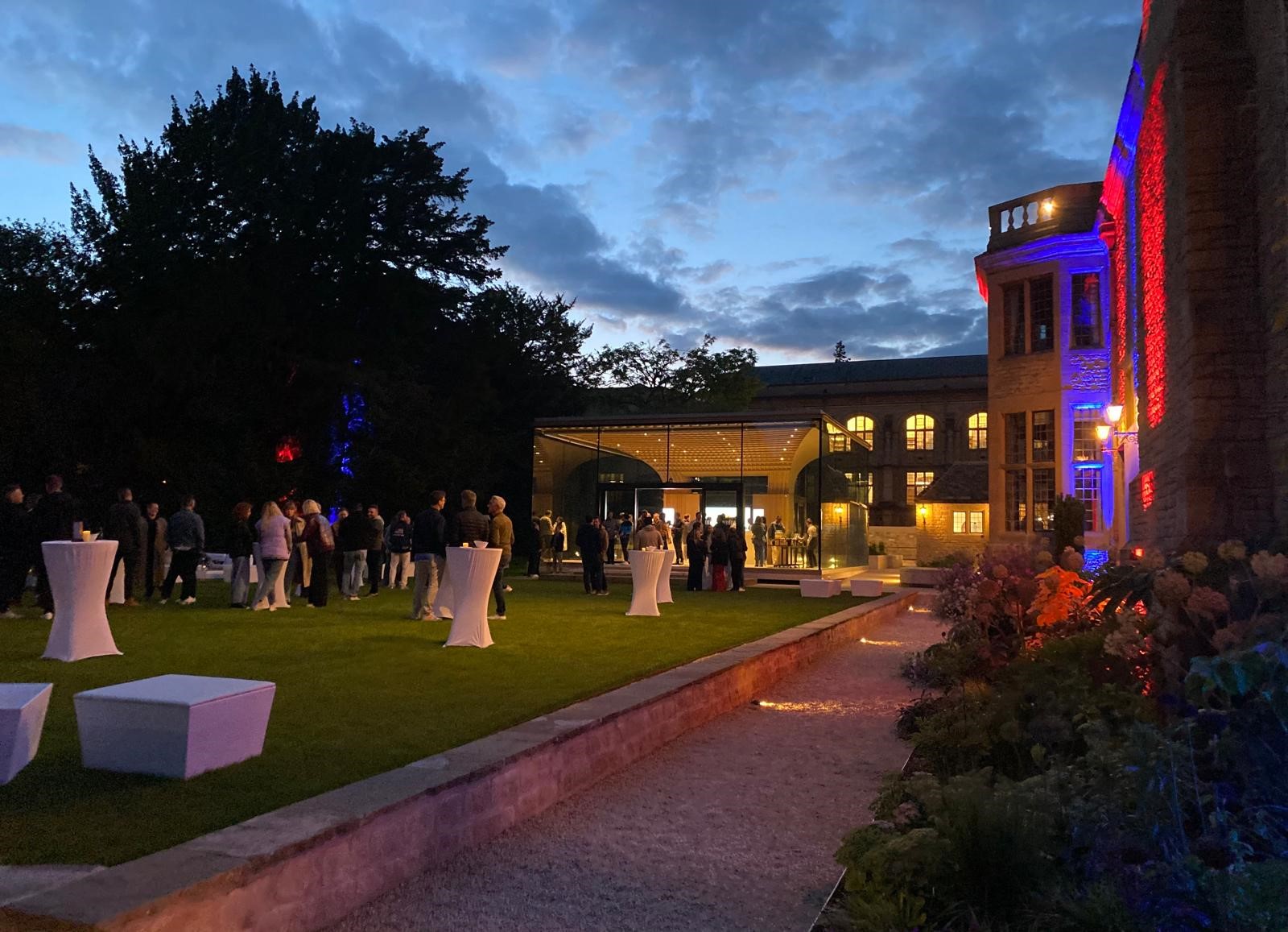 Drinks reception at night on the Rhodes House West Lawn with the Glass Pavilion in the background, poseur tables on the lawn, and red and blue uplighters on the outside walls
