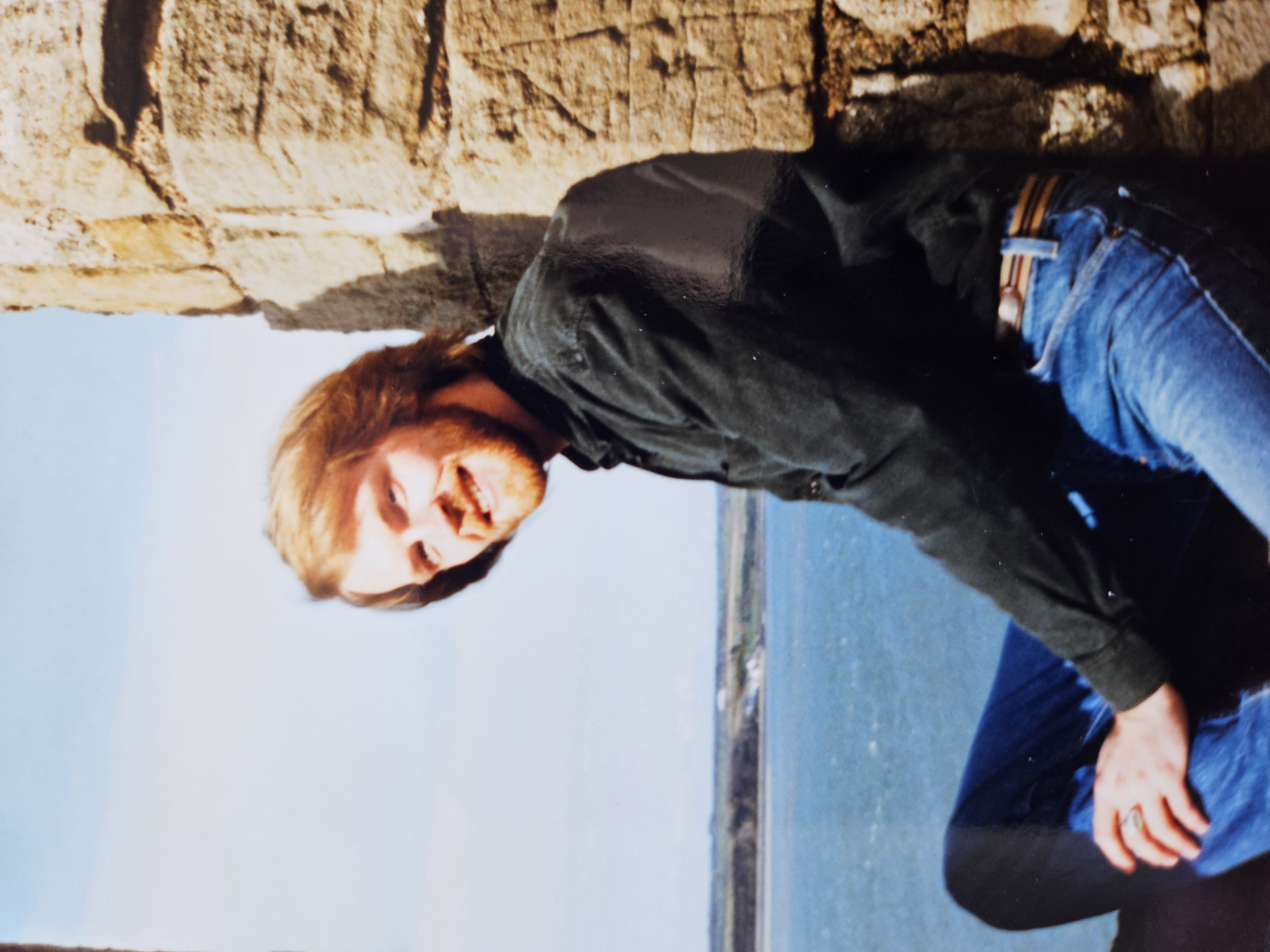 Eric sat on a stone structure wearing blue jeans and a shirt. He is looking into the distance, and a bright blue sea and sky is seen behind him.