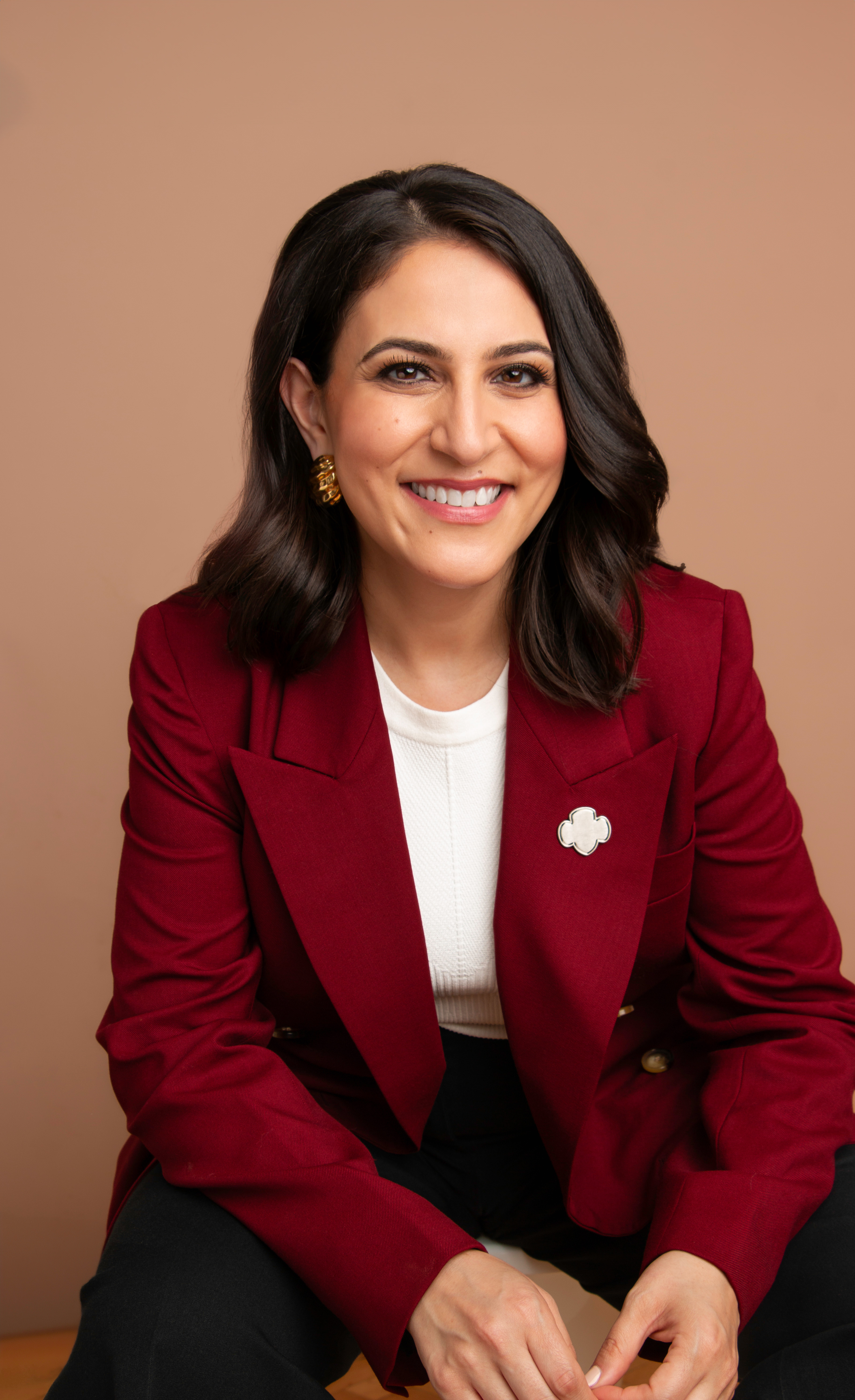 Portrait photo of Noorain wearing a  red blazer, leaning forward and smiling.