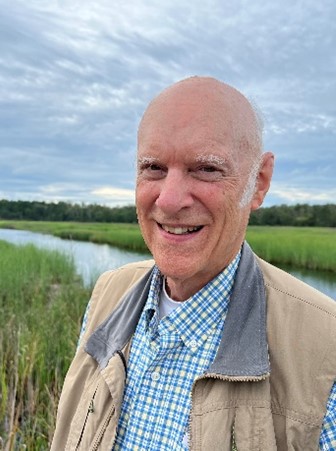 The picture shows Peter in a brown jacket and striped-blue shirt. There are grasslands in the background.