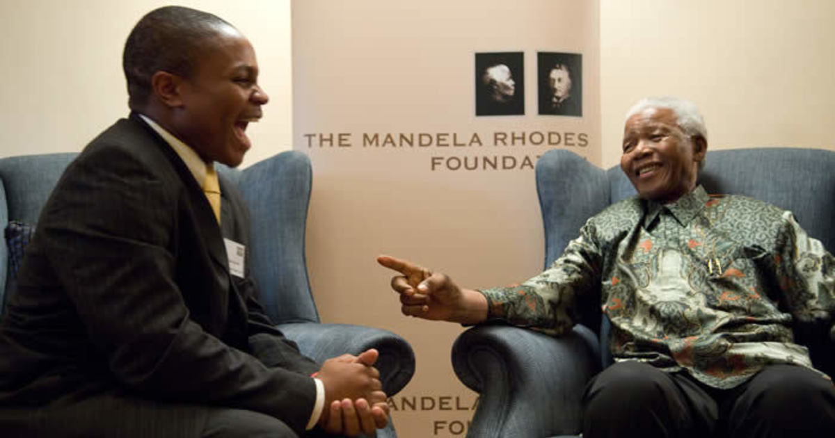 An image of Nelson Mandela sitting opposite a Rhodes Scholar. They are sitting in armchairs, both smiling and in the middle of a conversation. In the background is a banner that says 'The Mandela Rhodes Foundation'