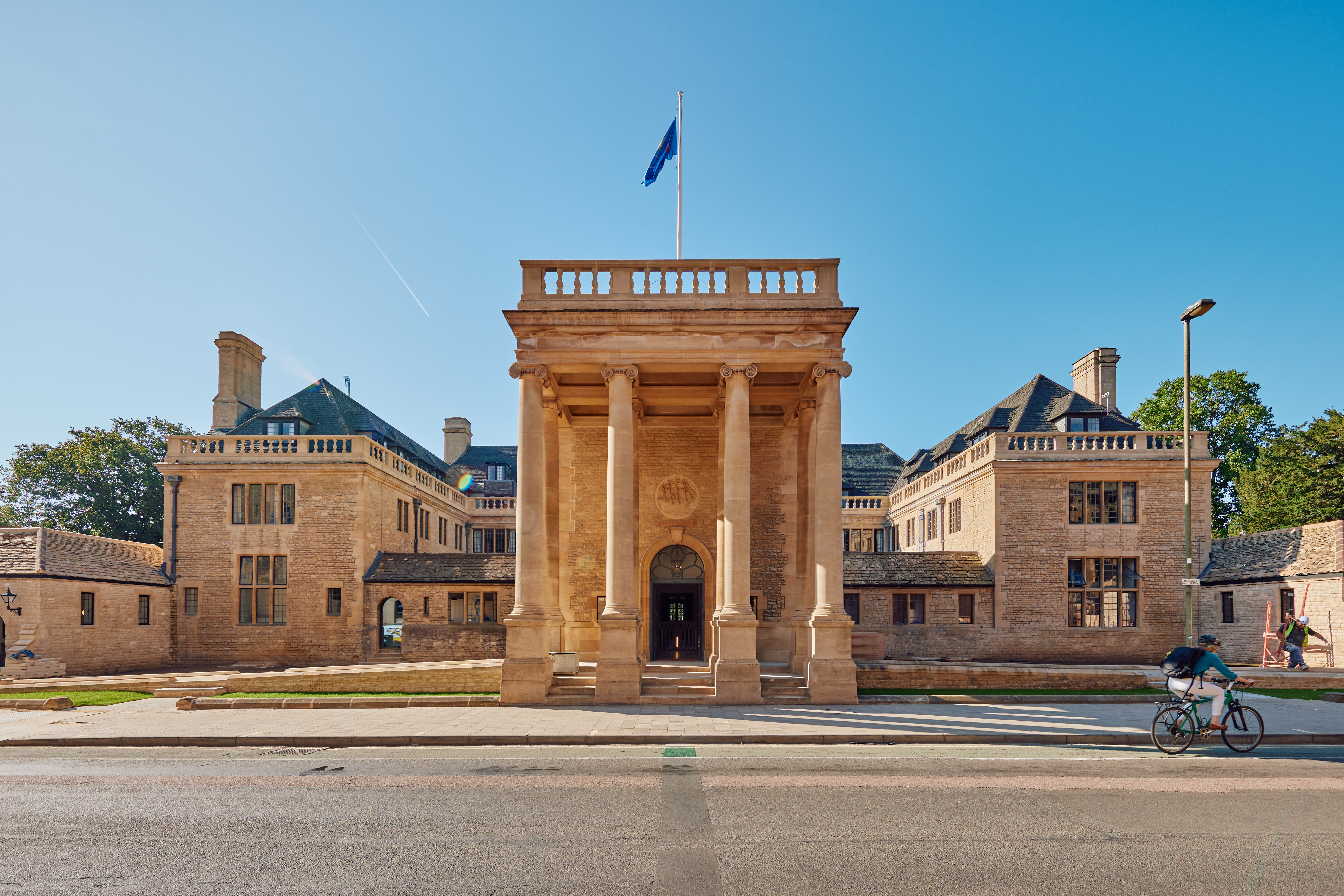 An Image of Rhodes House from the front. It is a stone building with four large pillars at the front and is illuminated by the sun under a clear blue sky. 