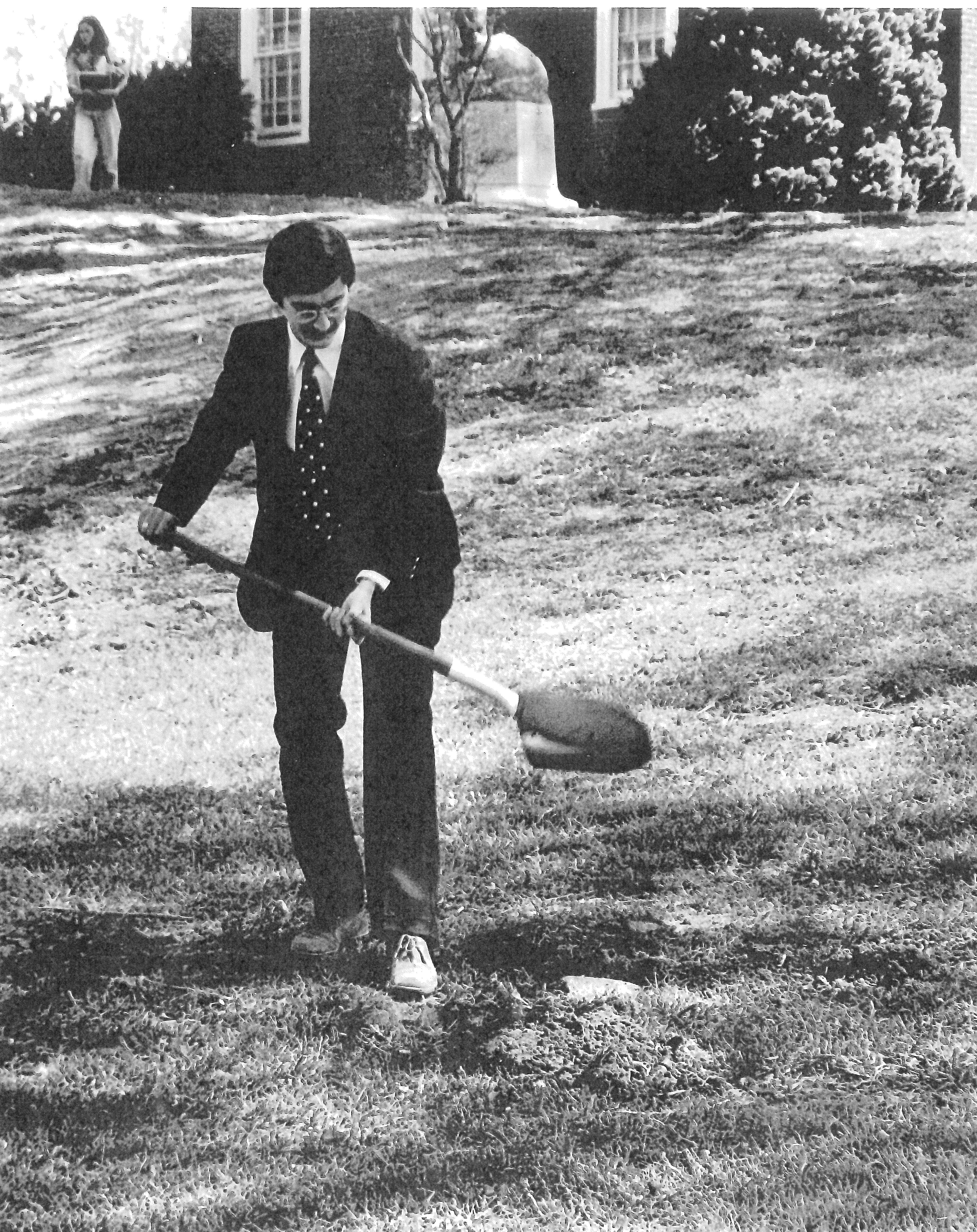 Black and white photograph of Larry in a suit digging into a hole in the ground. A brick home is seen behind him and a woman is walking in the background.
