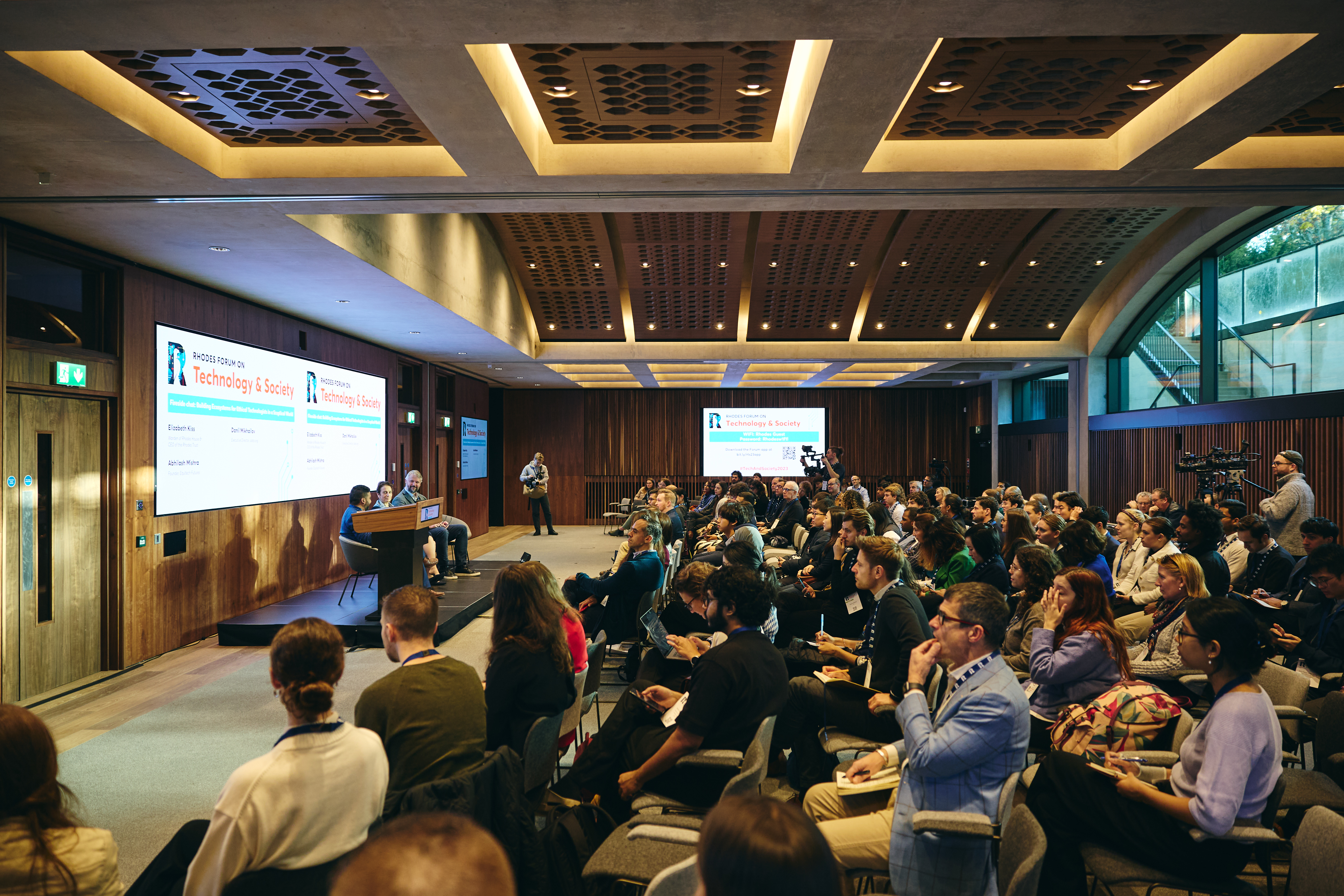An image of a conference room filled with people listening to three speakers on stage with two cameras filming.