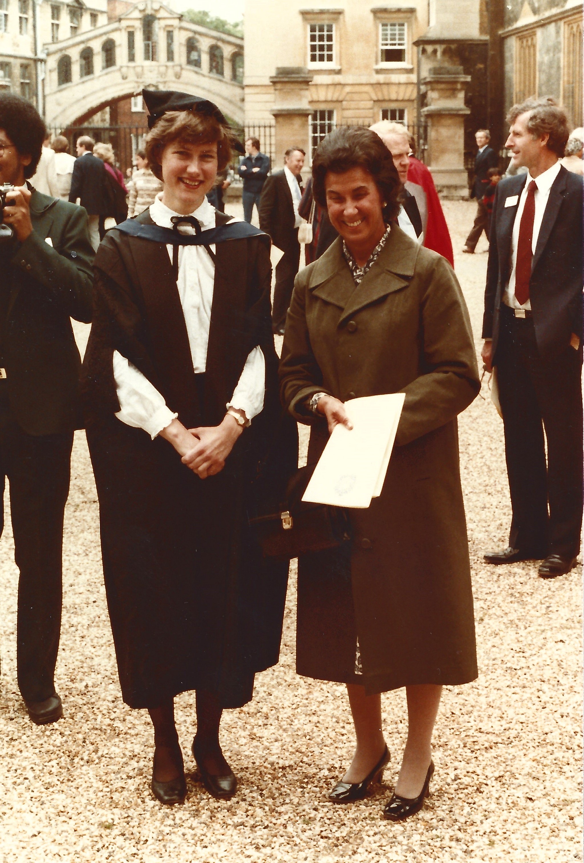 Margaret stood in graduation gown to the left of her mother, both smiling, with people walking behind them.