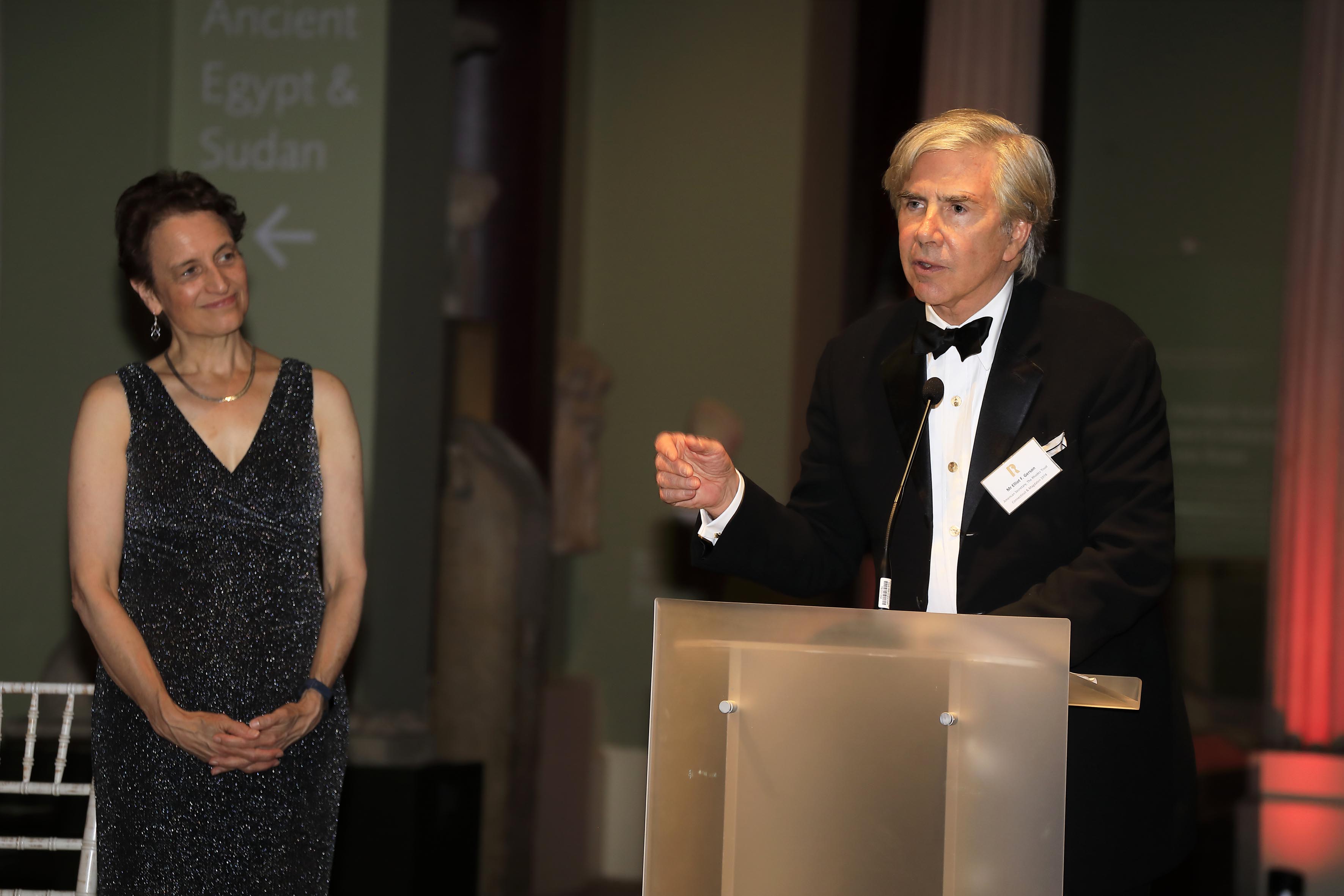 Elliot stood at a podium giving a talk, Elizabeth Kiss stood to the right and smiling.