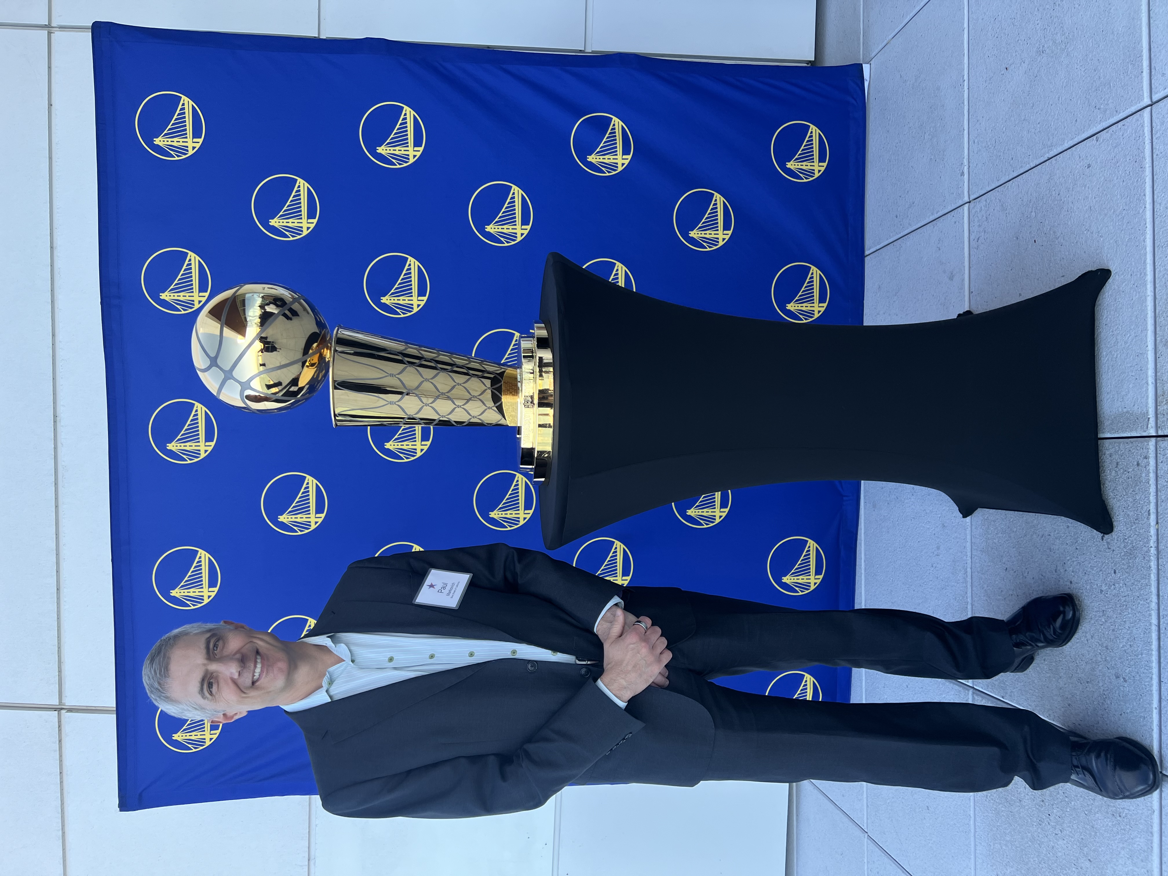 Paul stood in a suit posing next to a gold trophy which is placed on a black stand. The background is a blue display.