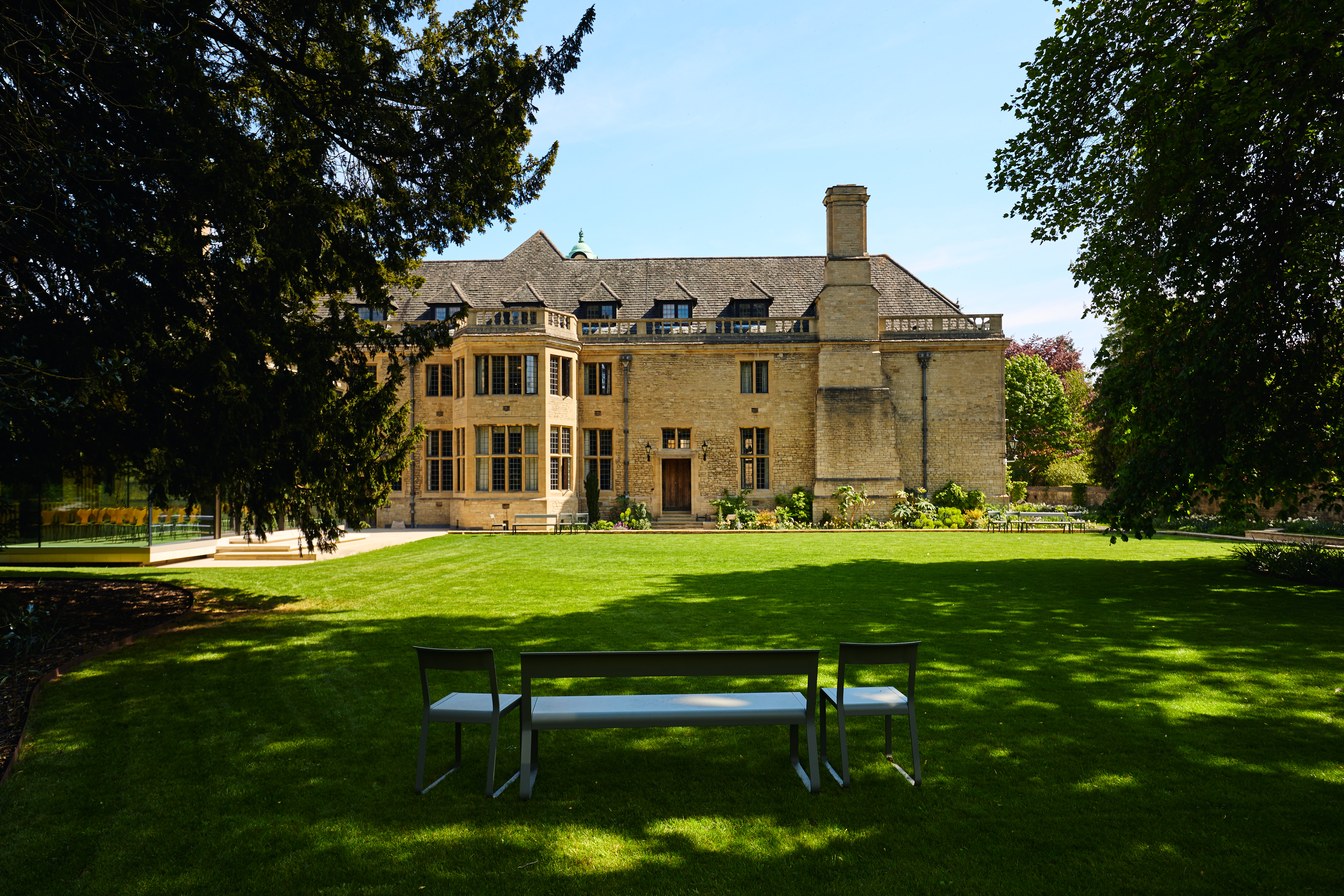 Daytime view of the west gardens with Rhodes House in the background and garden furniture in the foreground in the shade. 