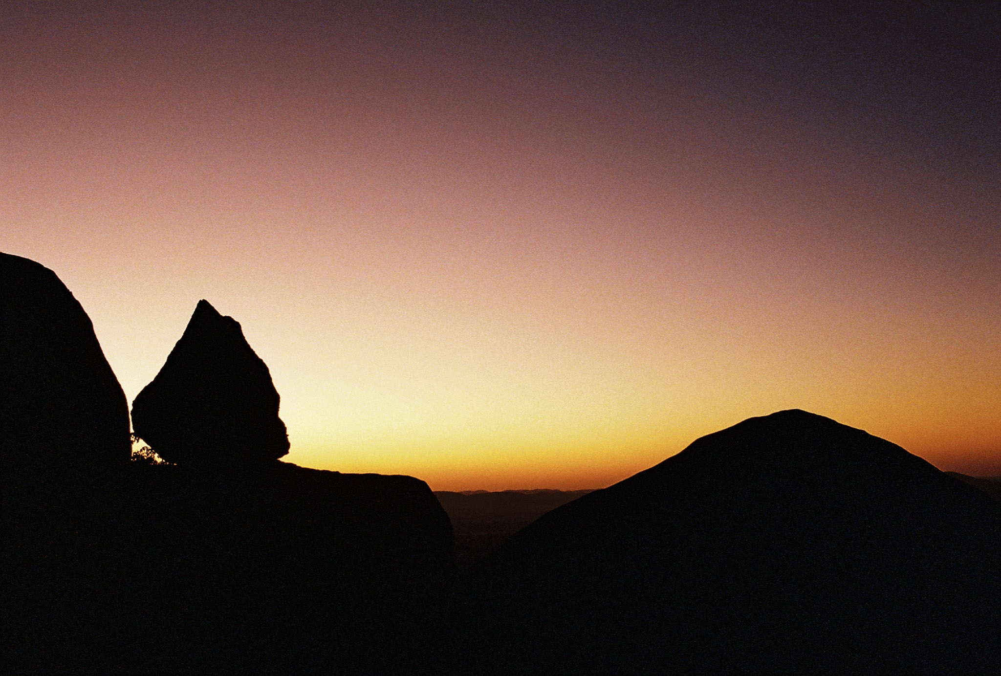 A purple-orange sunset horizon viewed from altitude atop the Great Zimbabwe Hilltop complex with a tear-drop shaped boulder foregrounded in silhouette delicately perched as if about to roll