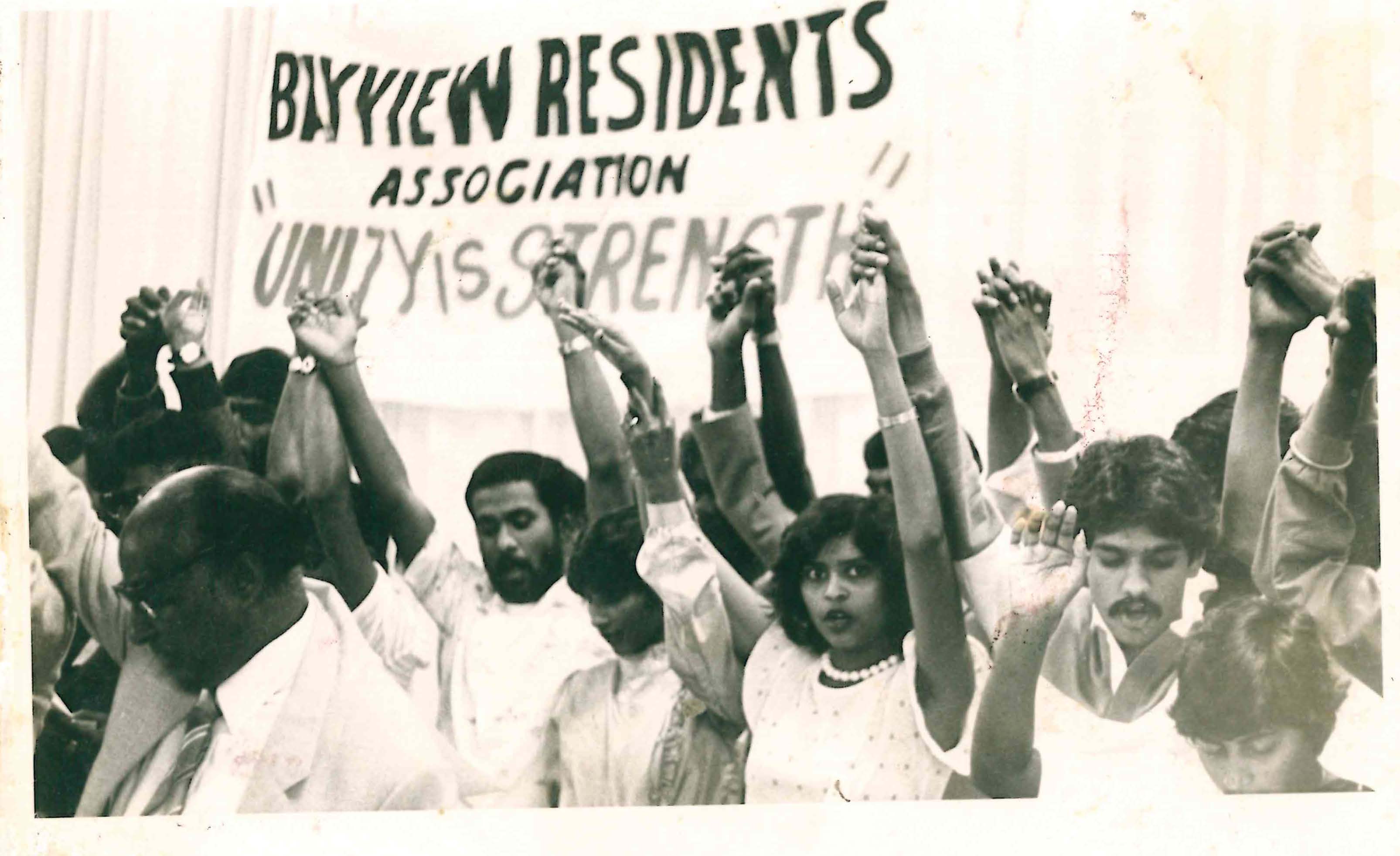The picture shows Kumi protesting with other people. Their hands are in the air and they hold a banner on which is written 'Unity is Strength'