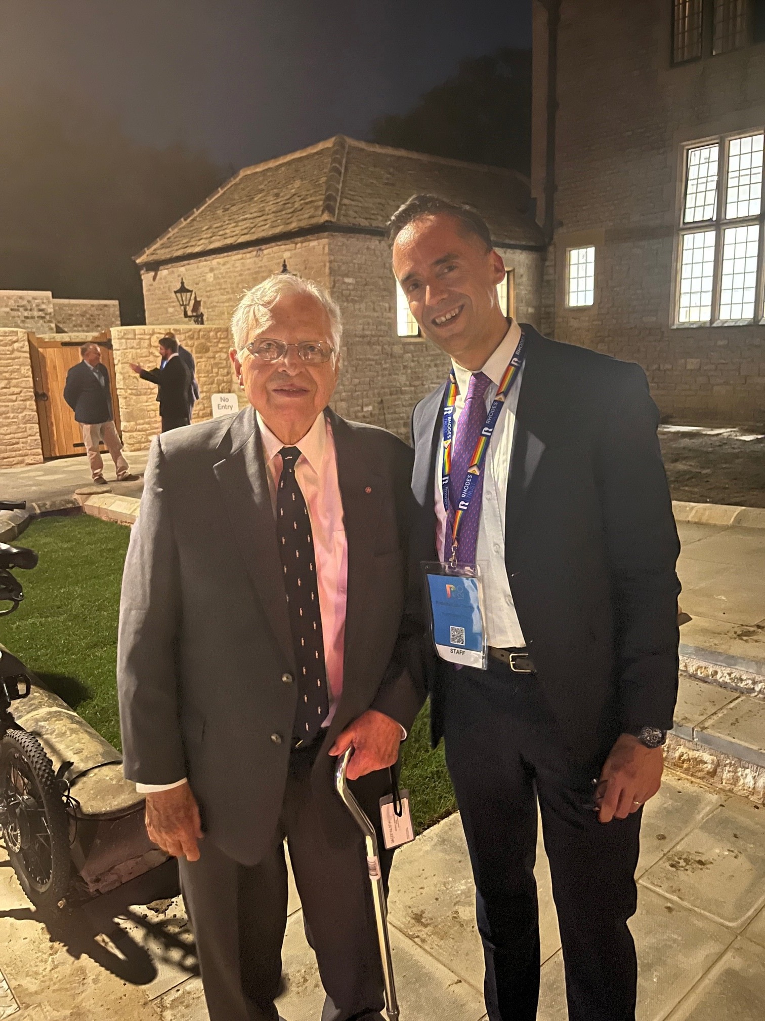 A picture showing John in a suit standing next to Rodolfo, also in a suit, with the Rhodes House in background