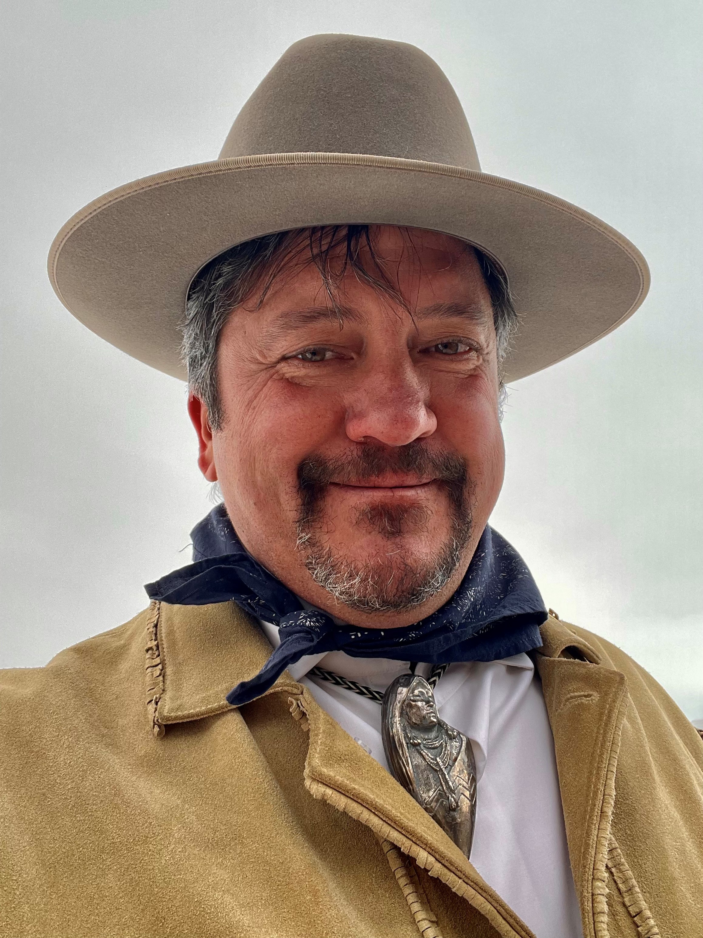 Deacon taking a selfie, wearing a tan coloured jacket and a hat, looking down at the camera and slightly smiling.