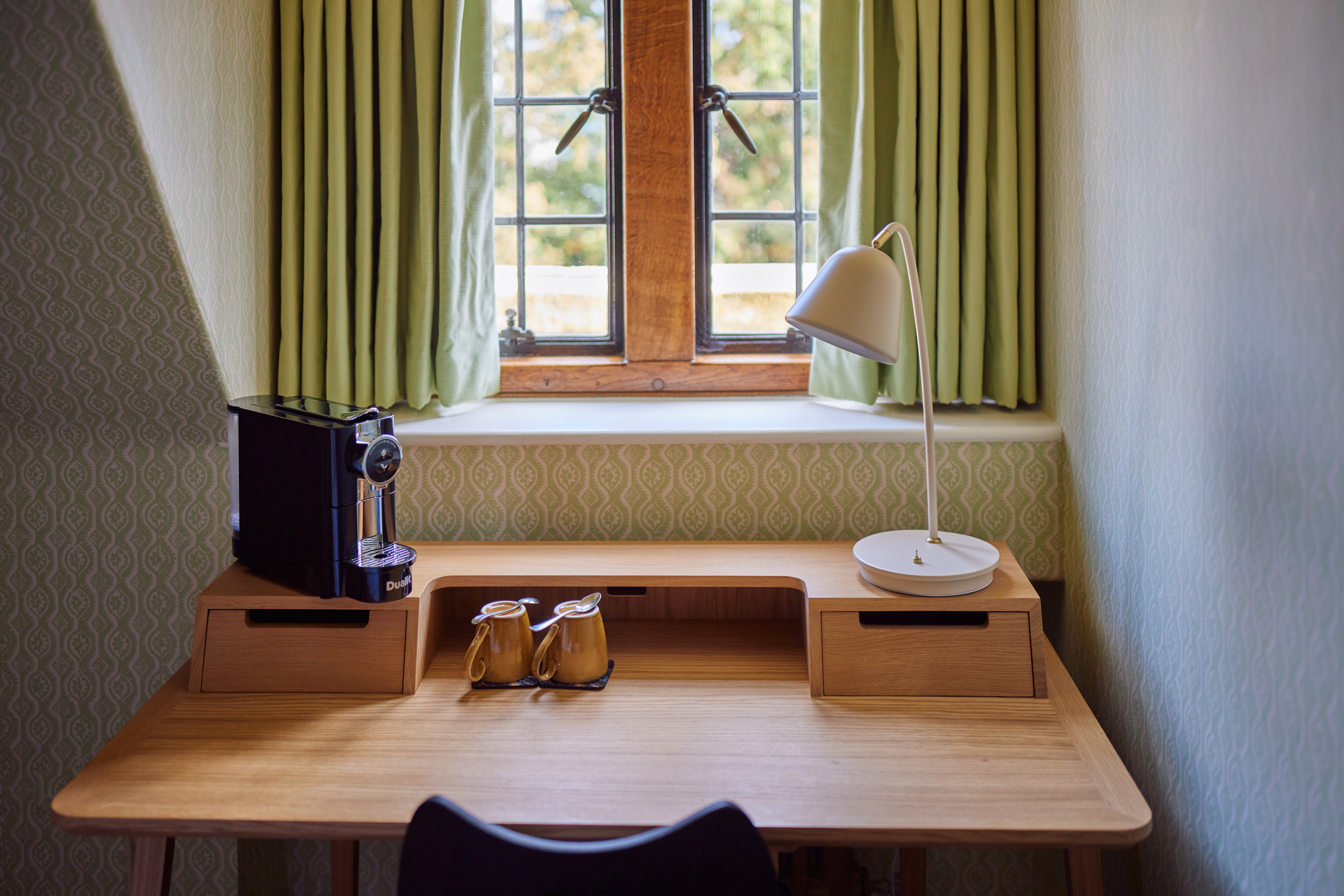 Rhodes House accommodation - A Wooden Framed Window With Green Curtains Sits Above A Wooden Desk; Sitting Below It The Desk Has A Coffee Machine Two Mugs And A White Lamp