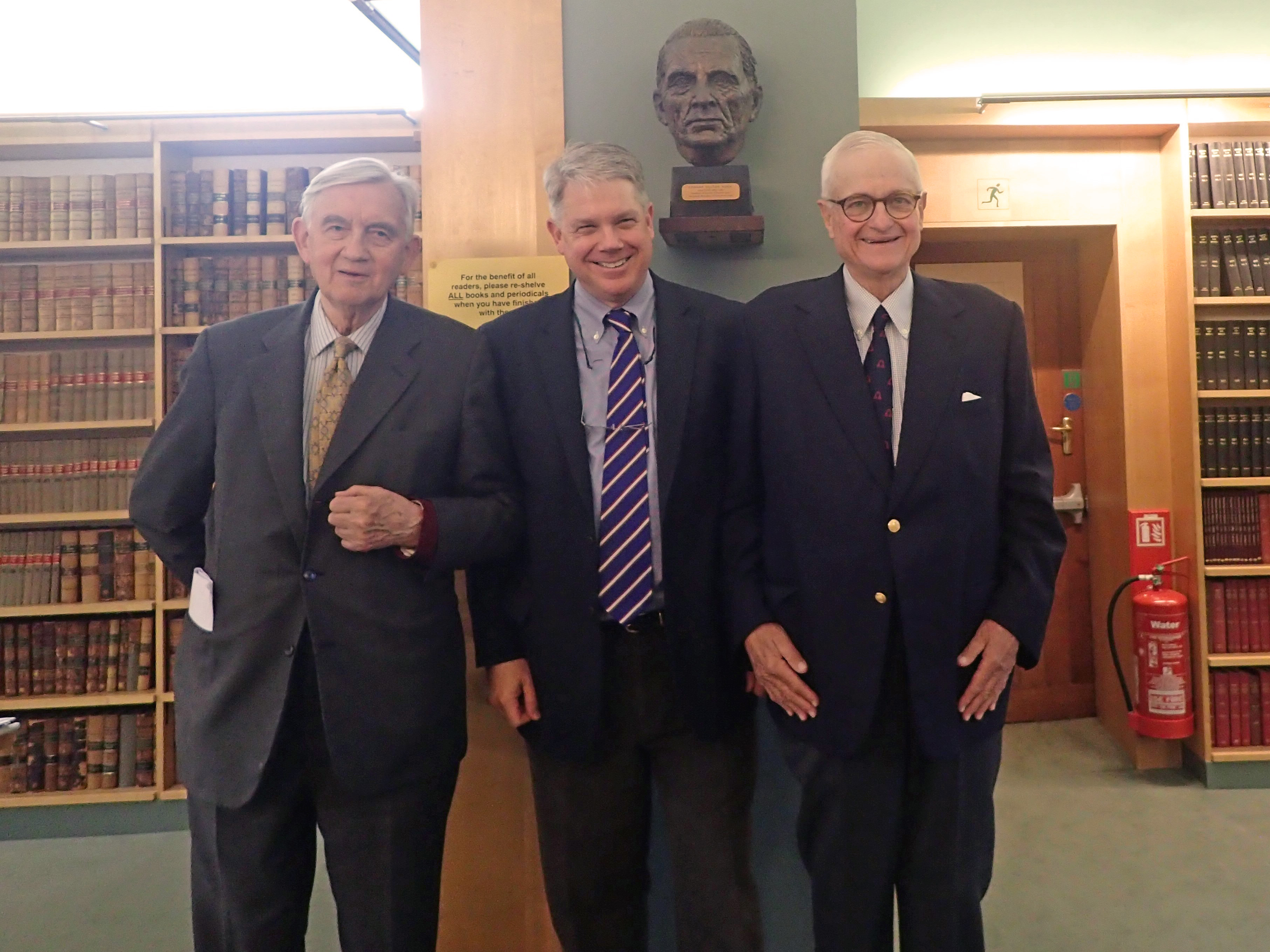 Three men stood in a library, wearing suits and smiling into camera.
