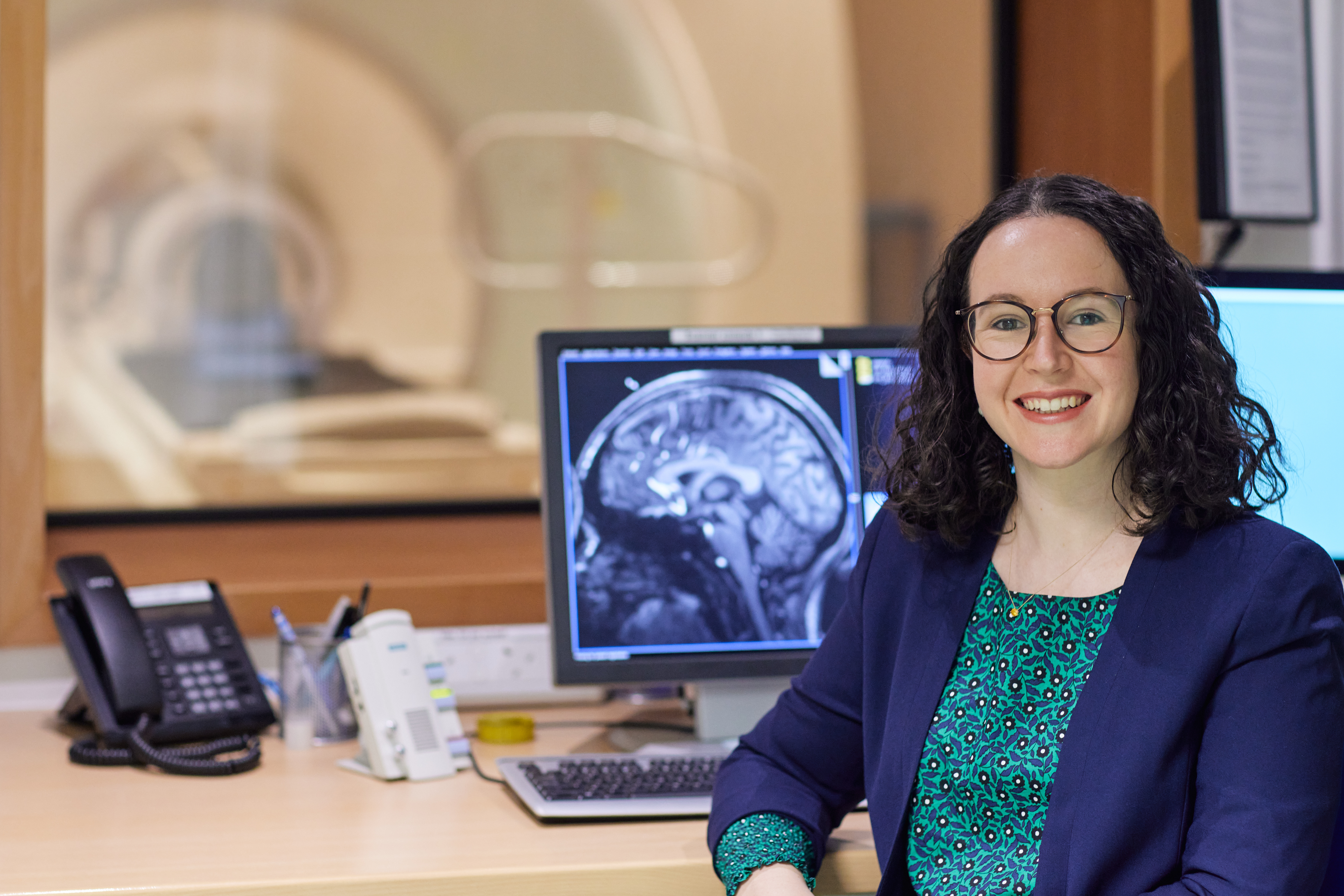 A woman smiling at the camera against the backdrop of a medical scanner with a brain scan on a screen behind