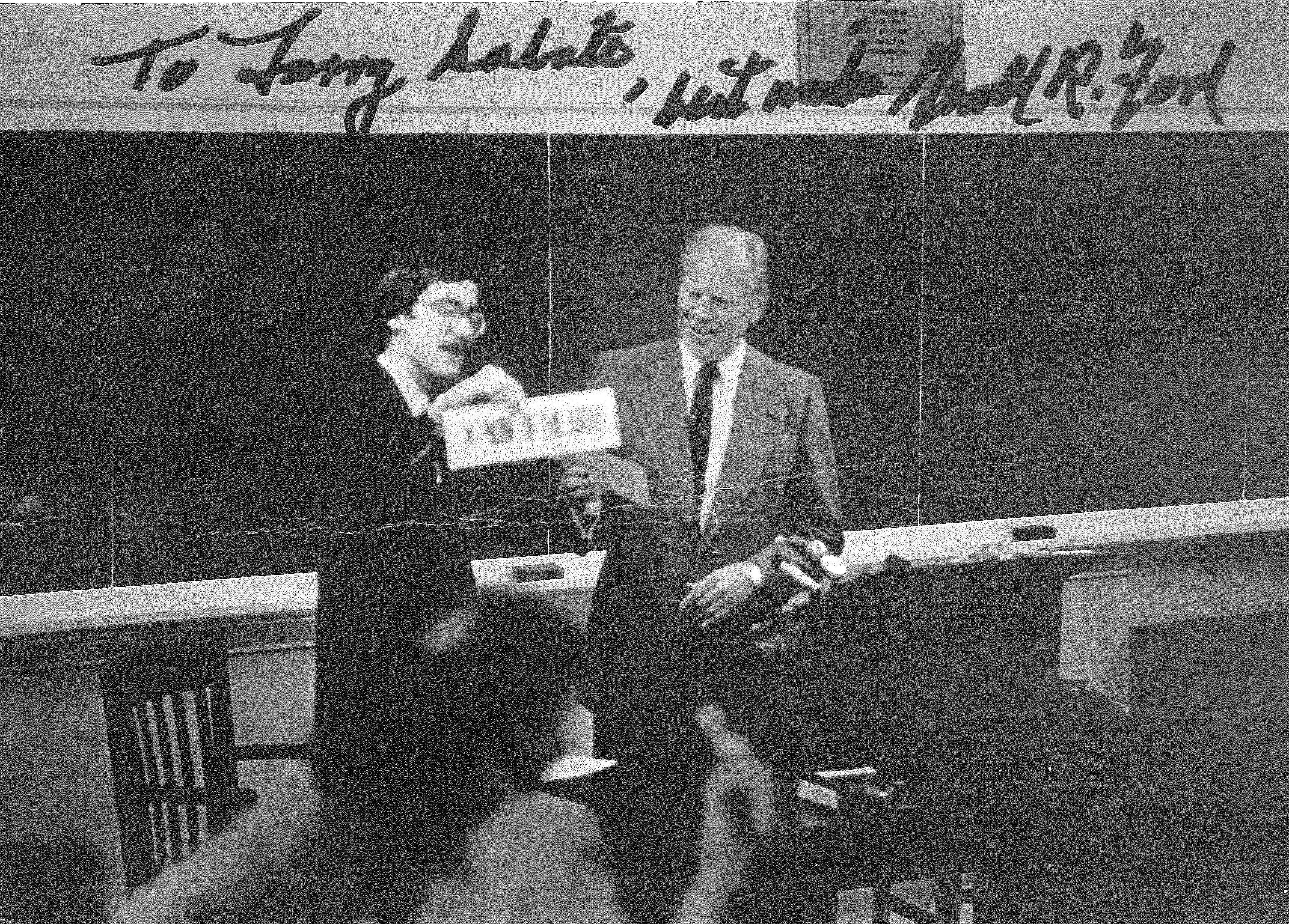 Black and white photo of Larry Sabato and Carter. Sabato is handing Ford a piece of paper, but the writing is illegible. There is handwritten text at the top of the photograph which reads 'To Larry Sabato, best wishes Ford'.
