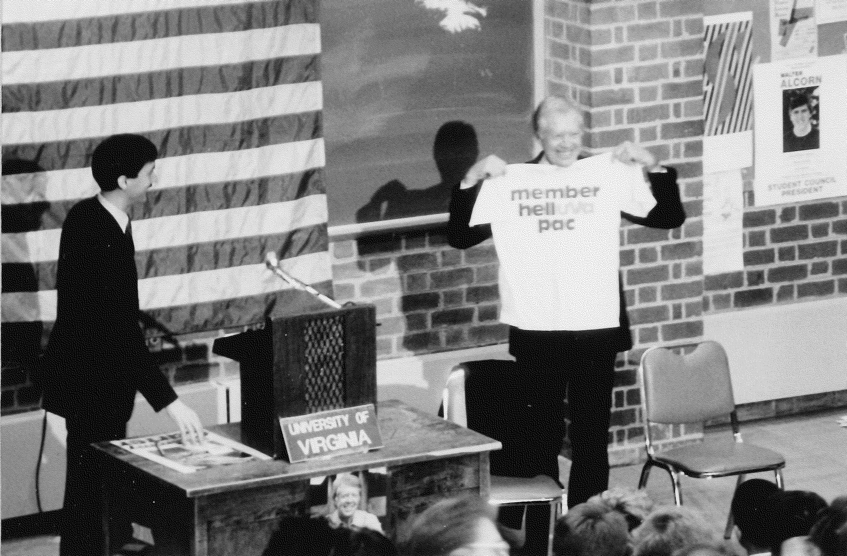Black and white photograph picturing Larry and Carter. They are before a crowd and by a desk with a sign that reads 'University of Virginia'. The US flag is behind them and Carter is holding a t shirt that says 'Member hell pac' (some text illegible) and smiling.