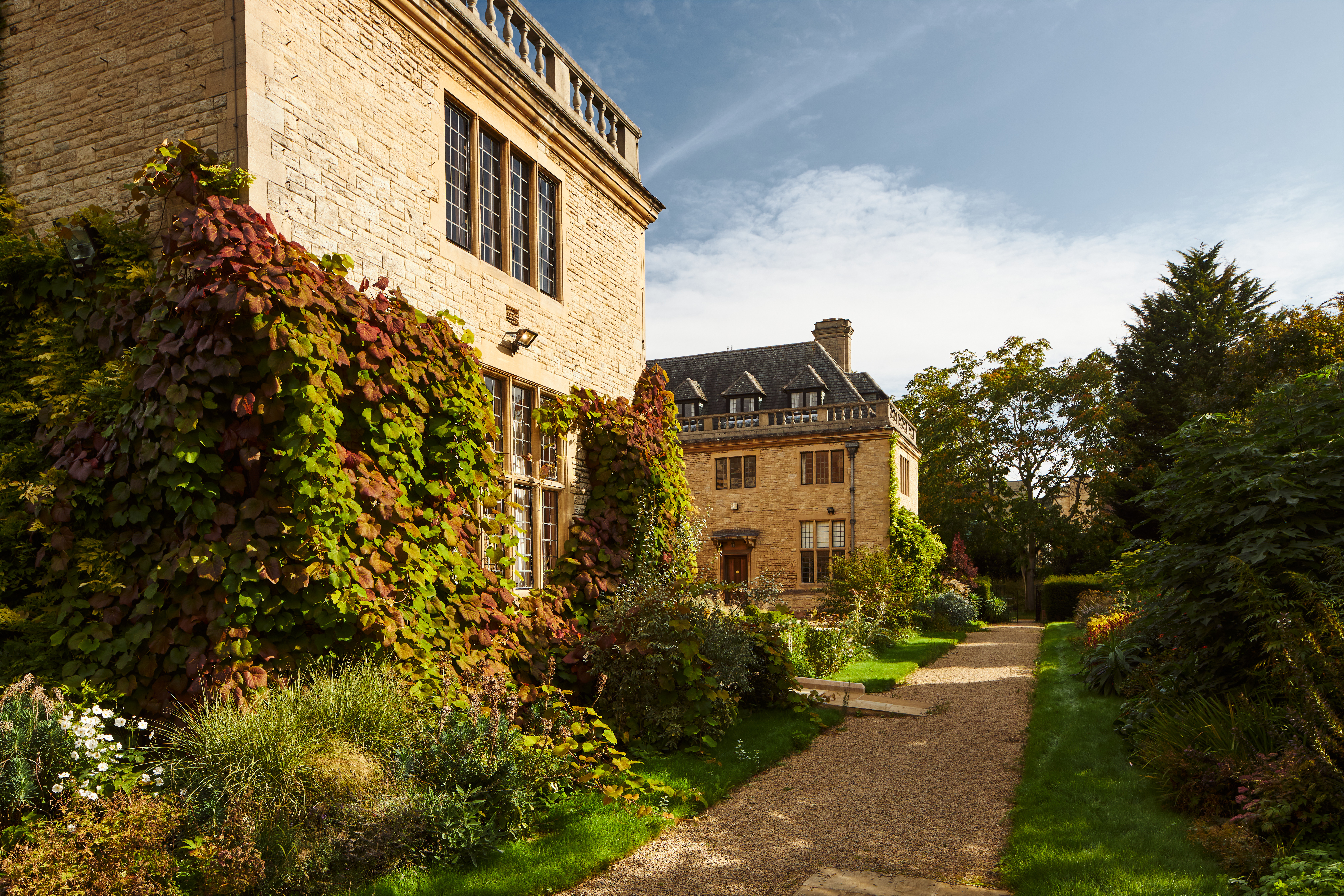 An image of the exterior of Rhodes House on a sunny day. The stone walls are covered in ivy and climbing plants, and there is a gravel path through a garden lined with hedges.