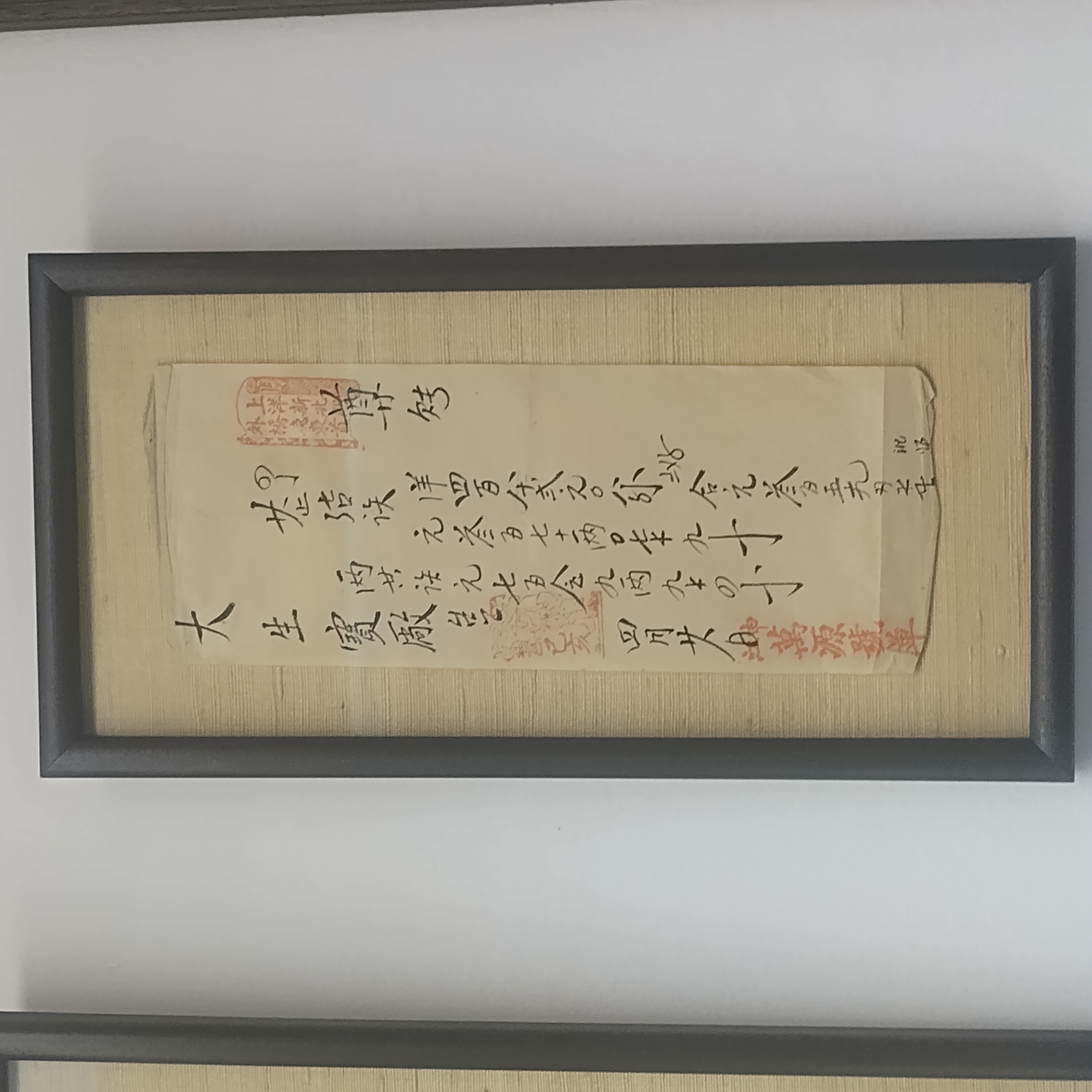 Photo is of a framed piece of paper, which appears to be very ancient and has handdrawn black Chinese characters with a few red prints.