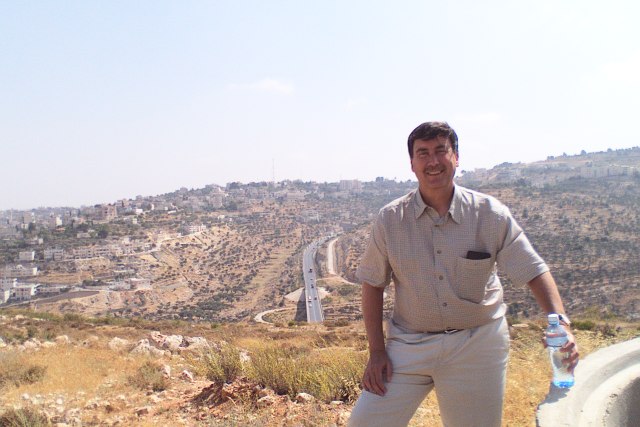 Sabato is pictured standing on the top of the hill. Behind him we see large buildings, grassy fields, and a motorway running through the middle.
