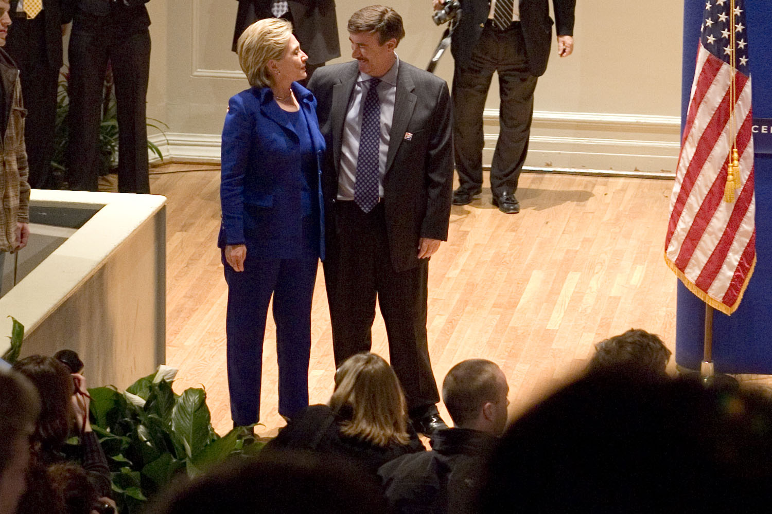 Hillary stood to the left of Larry in a blue suit. He has his arm around her and they are mid-conversation and smiling. They appear to be stood before a stage, in a room surrounding by people. The US flag is to the right.