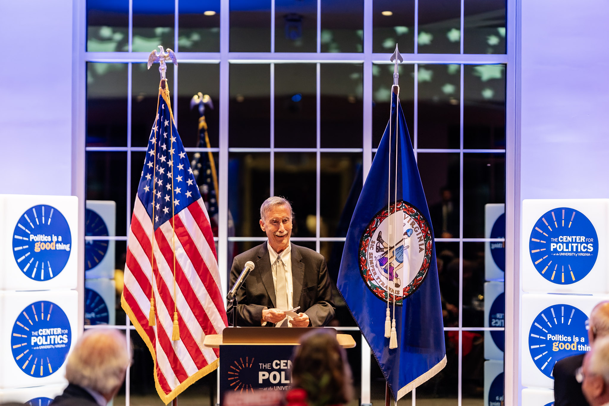 Larry stood before a crowd and behind a podium. The US flag is to his left, and the Virginia flag to his right. Banners around him read 'The Center for Politics'.