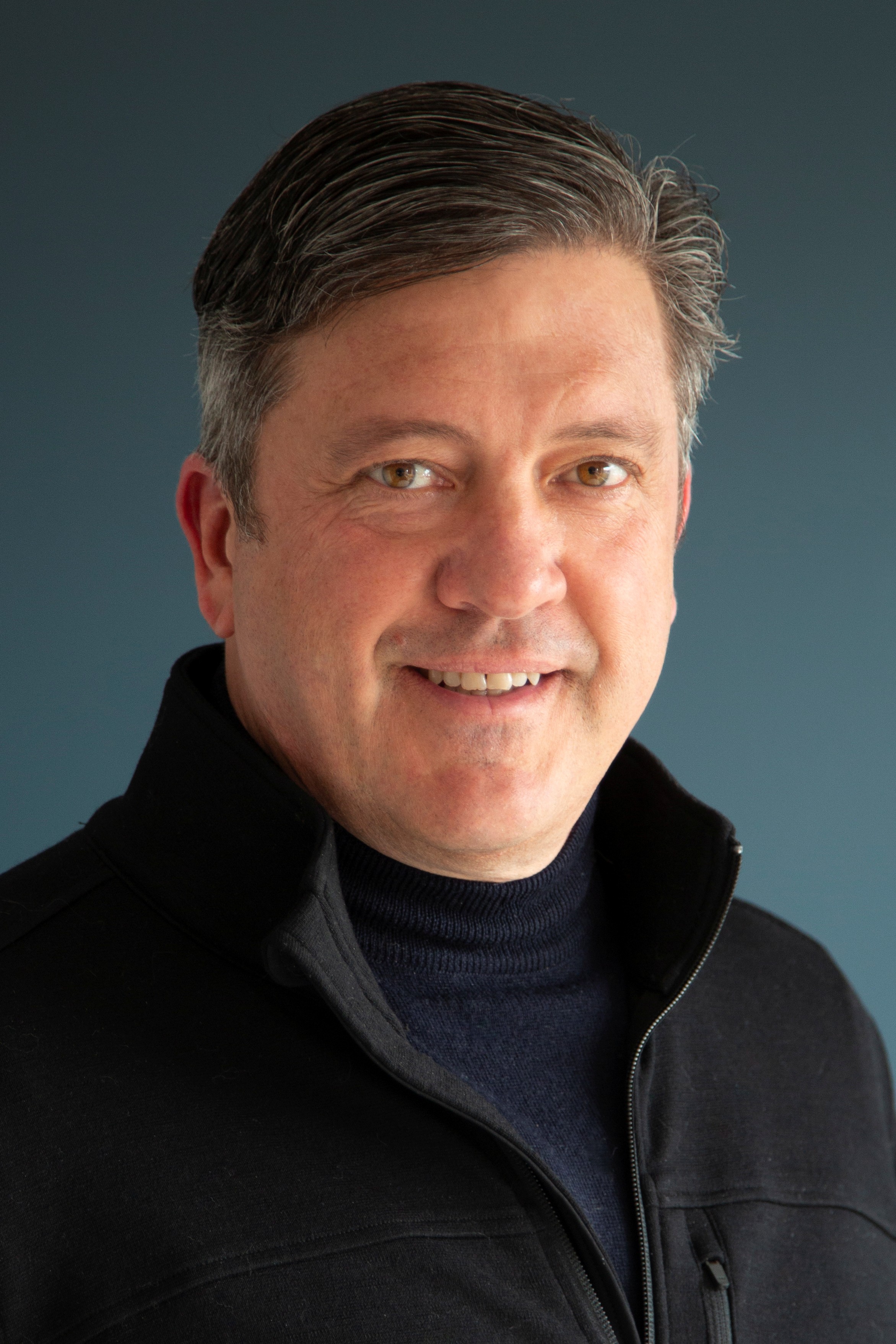 Portrait of Deacon wearing an alll black outfit, smiling before a blue background.