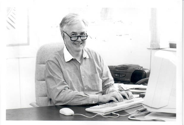 A black-and-white picture of Norton at a table, sitting in front of his computer, his fingers on the keyboard.
