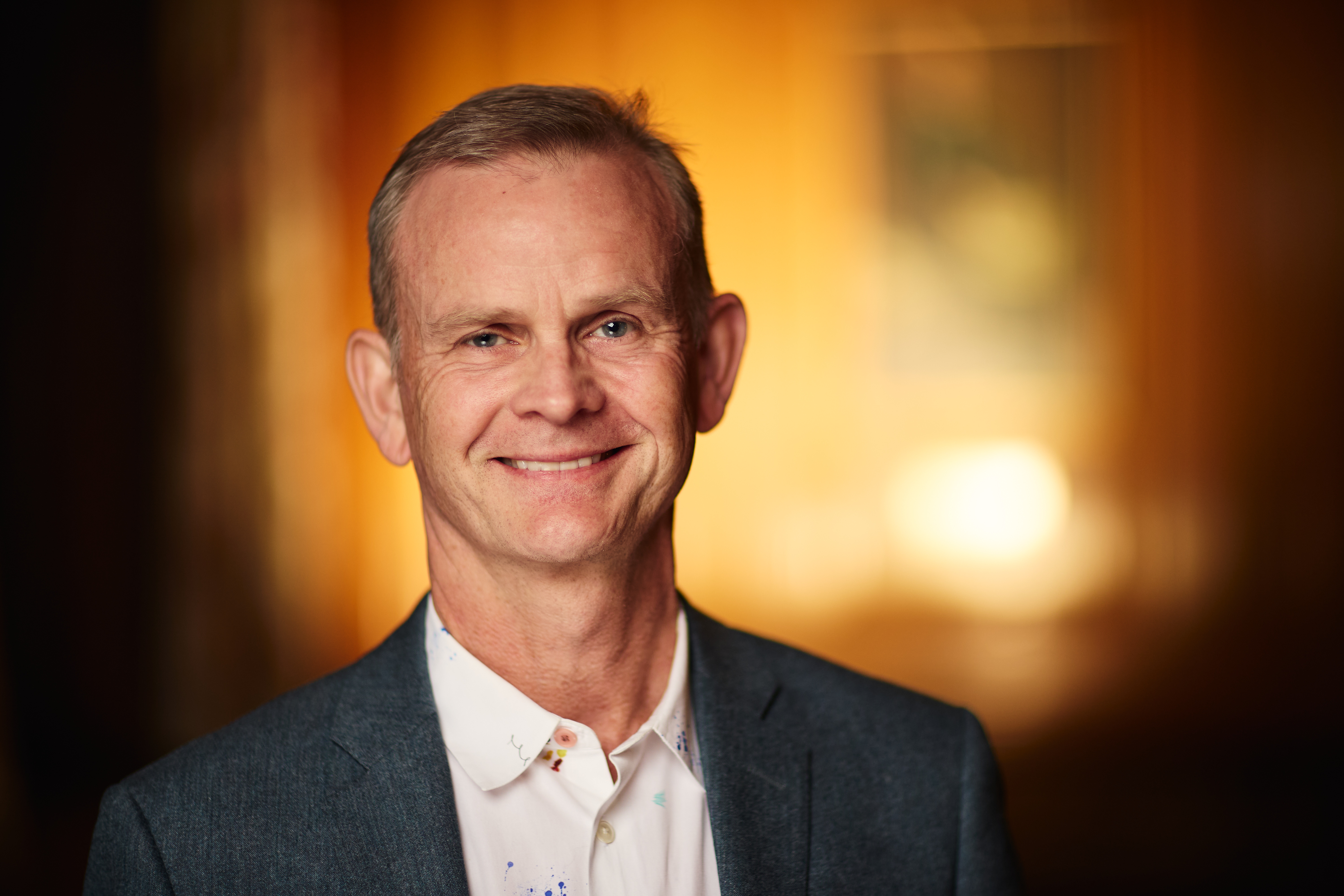 Headshot of Mark, in a white shirt and grey coat. He is smiling.