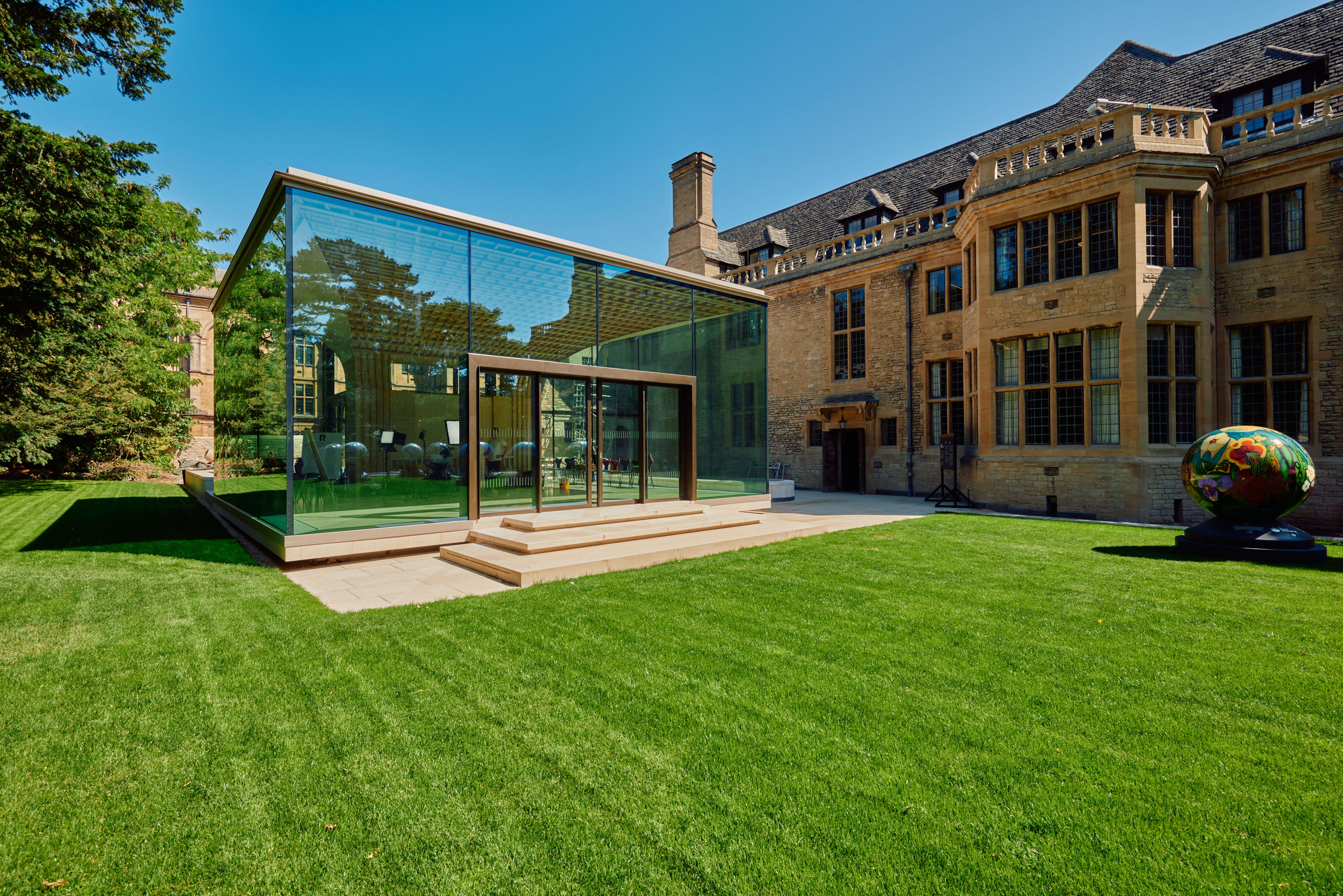A view of the Rhodes House Glass Pavilion, lawn, and west wall of Rhodes House in bright sunshine.
