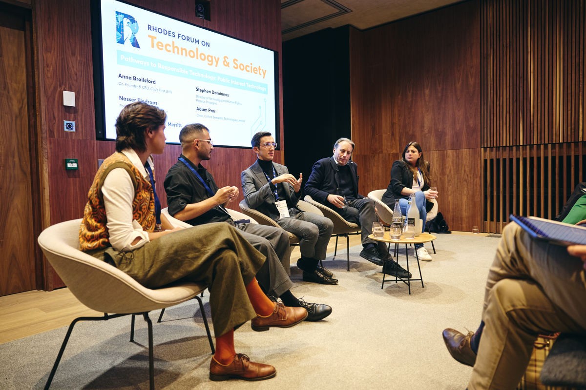 An image of 5 speakers sitting in a row at a conference in front of a crowd. Behind them a screen reads "Rhodes Forum on Technology and Society"