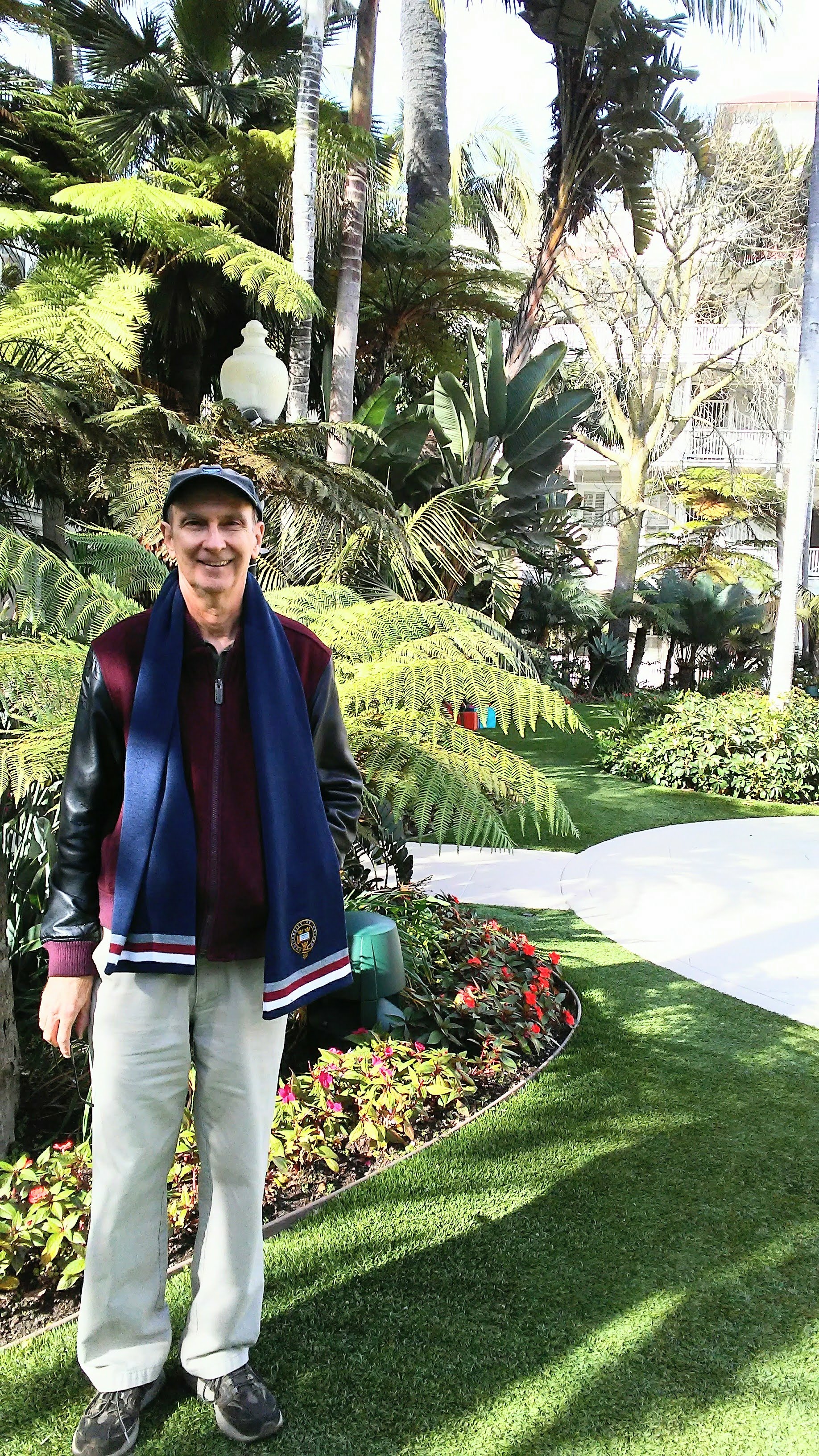 Roger stood smiling before a garden landscape with green grass, flowers, and many trees behind him.