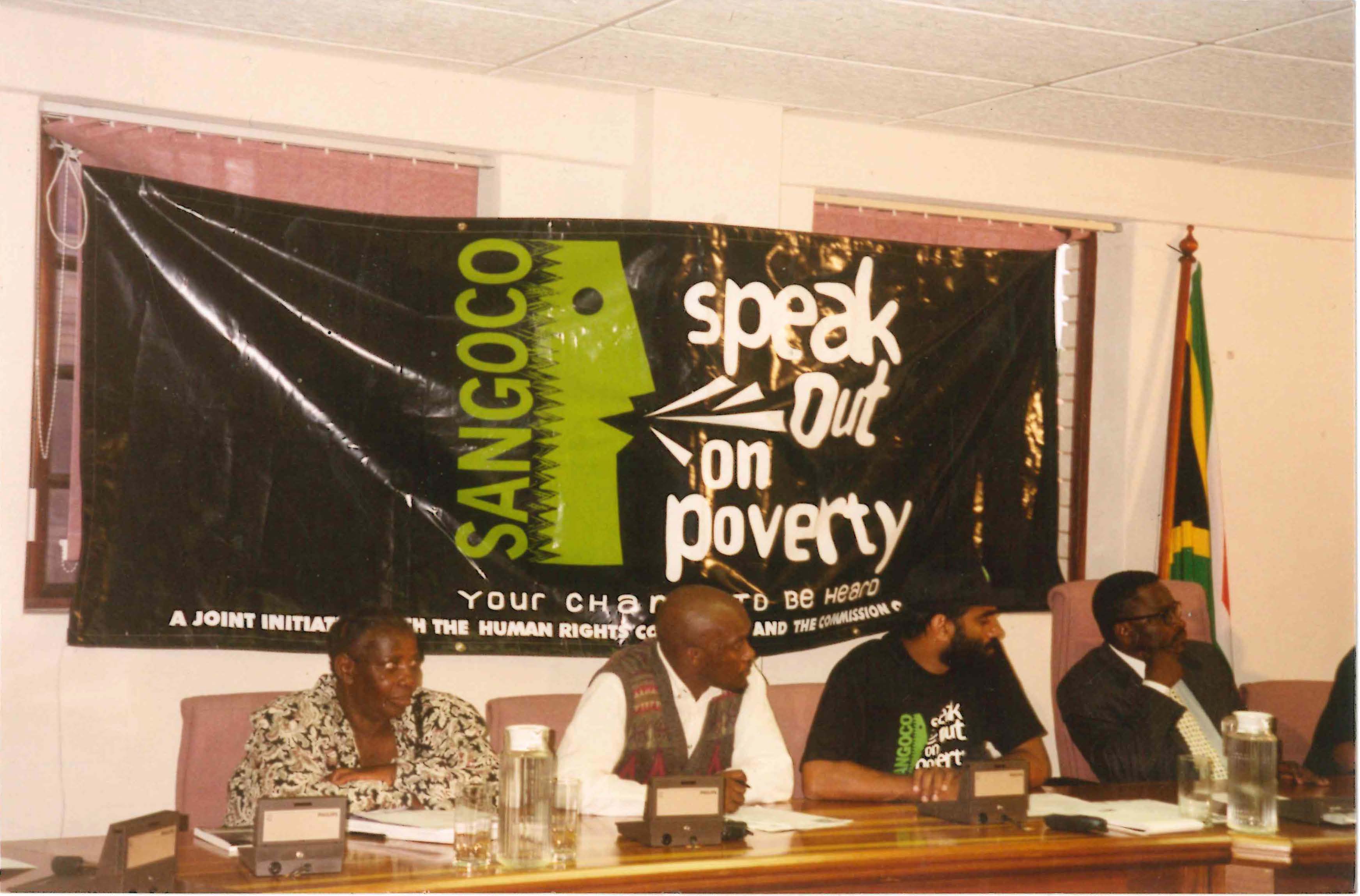 The picture shows Kumi speaking on a panel. The banner behind them reads 'Speak out on poverty'.