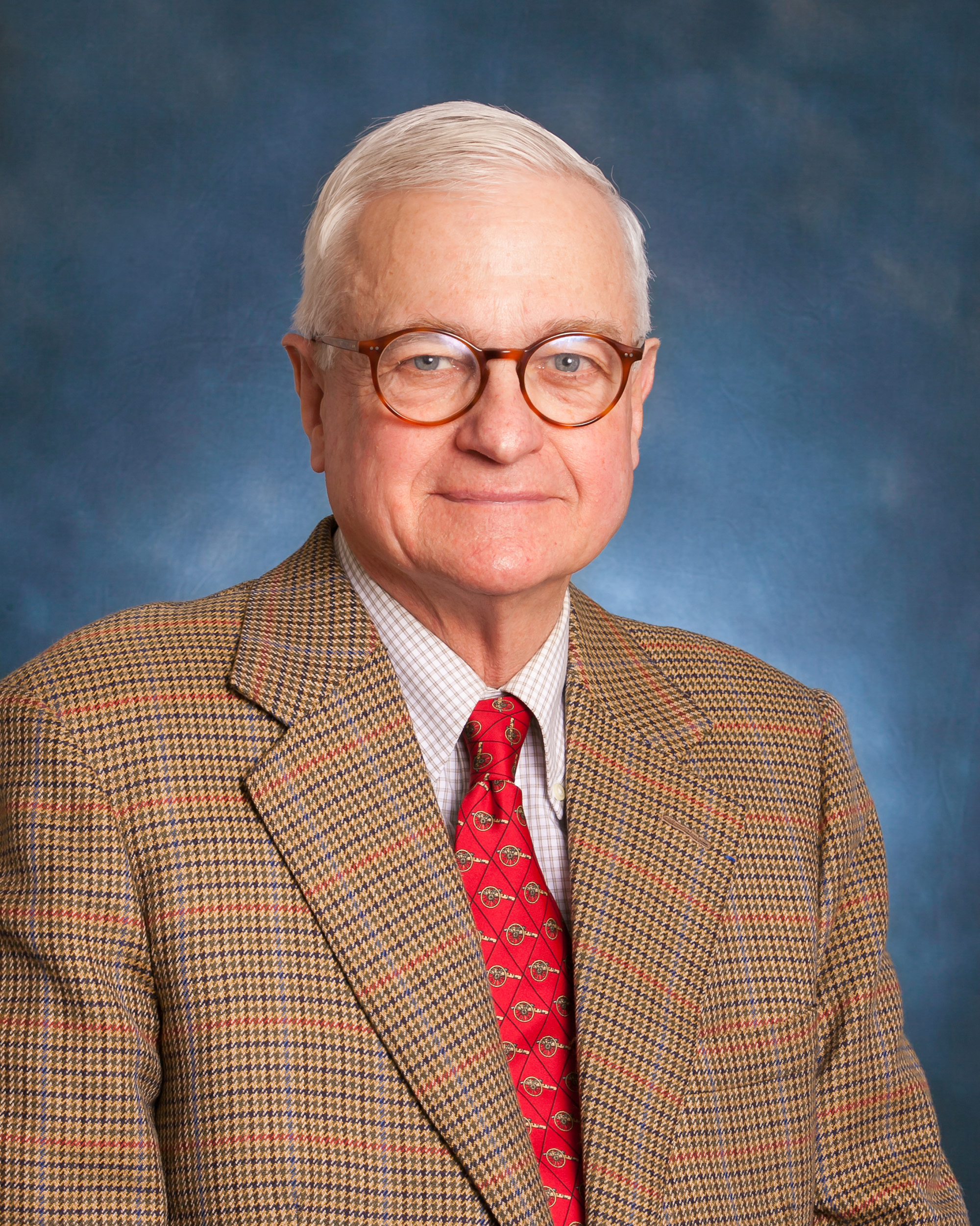 Portrait photo of Peter wearing a tweed suit, red tie, glasses, and a blue background.