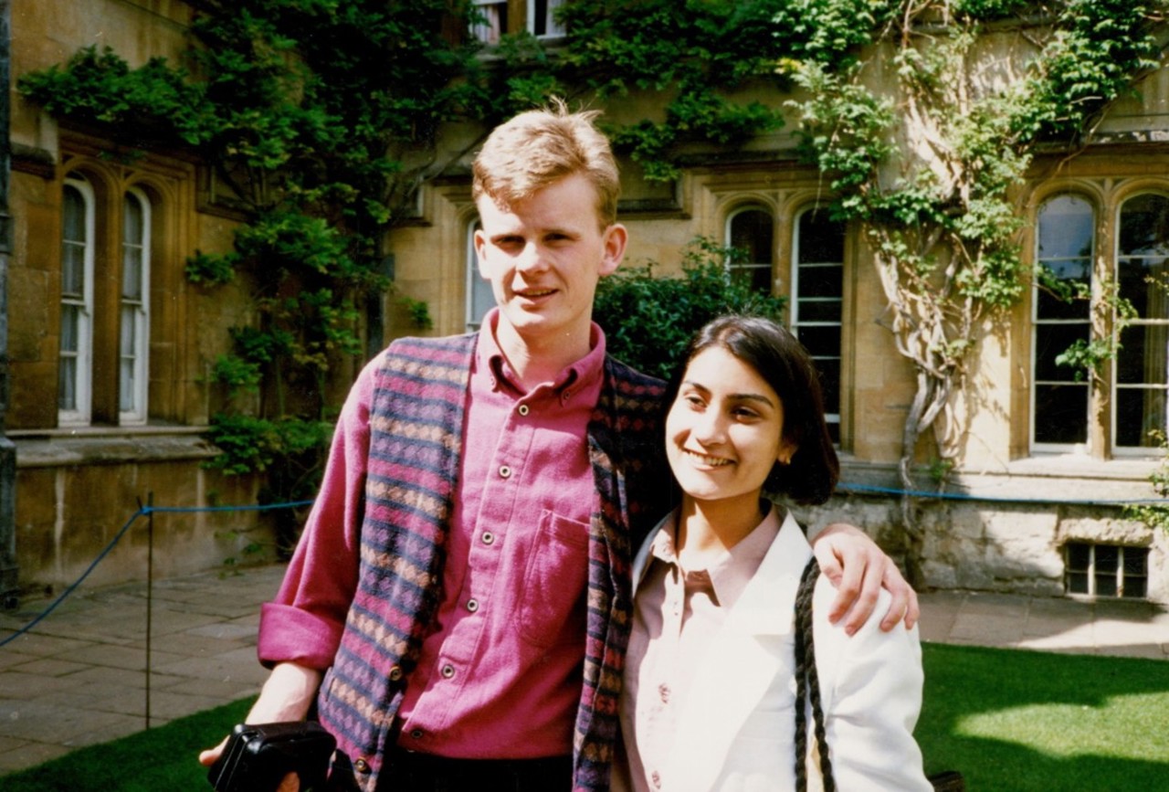 Mark. in a maroon shirt, standing with his hand around Shazia who is wearing a white shirt. The sandstone walls of University College buildings are in the background, with green creepers