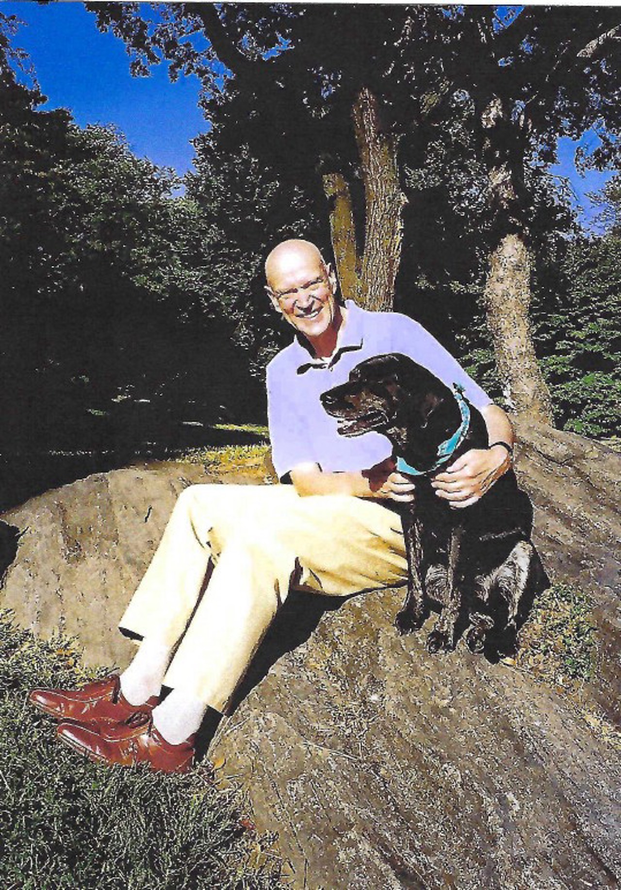 Willie sat on the ground, with a tree and nature behind him. He is holding a black dog and smiling into the camera.