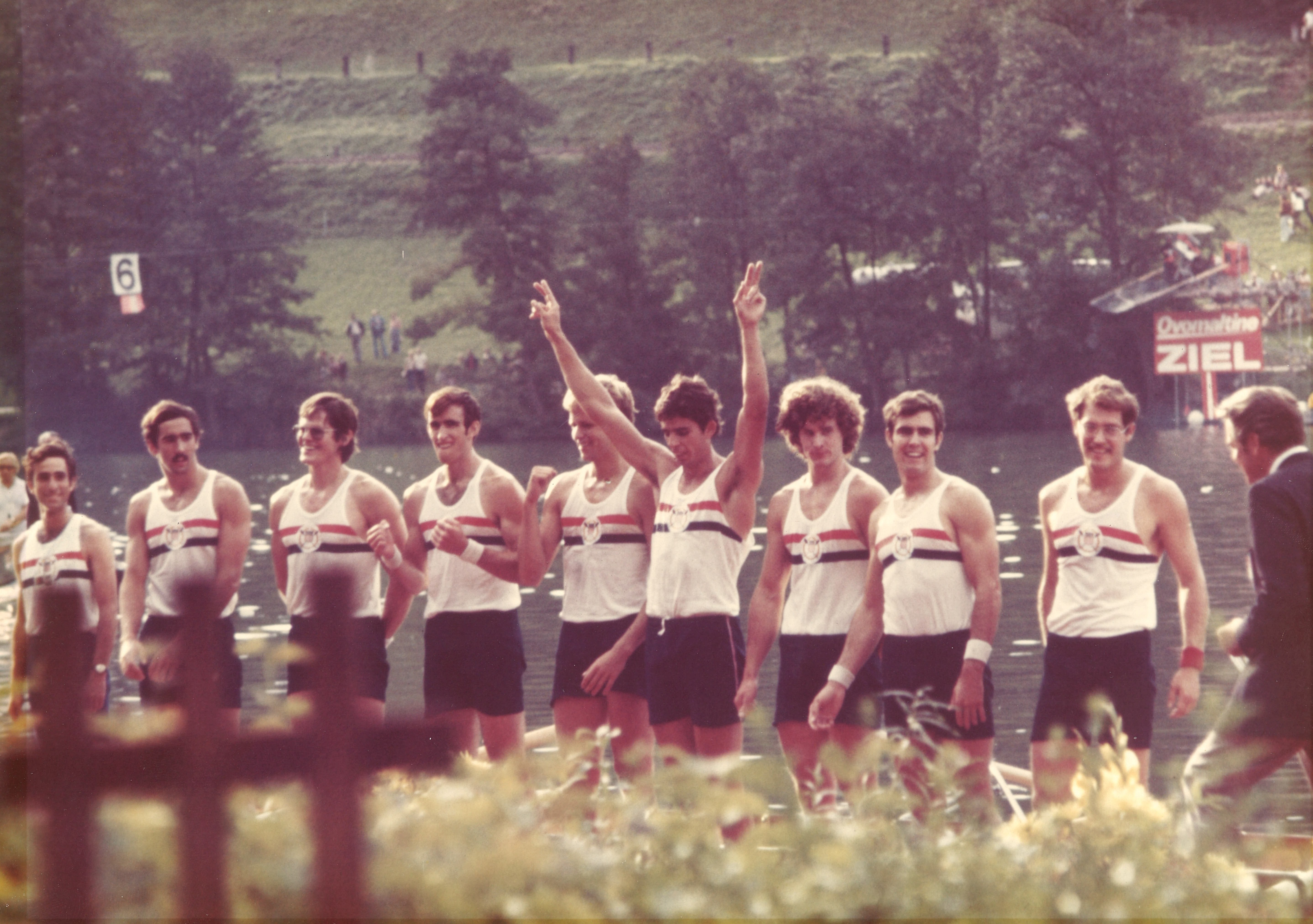 9 students are stood in a row before the water. They are all wearing rowing uniform.