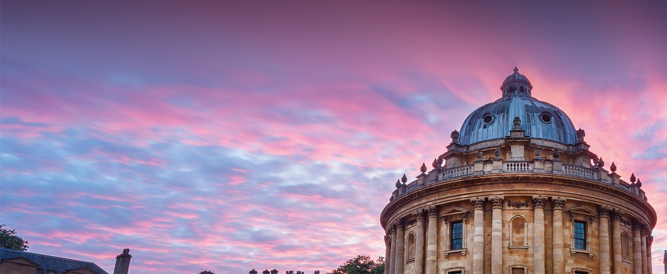 An image showing the top of the Oxford Radcliffe Camera building, made of light stone, with a domed roof. It is set against a sunset showing a blue and pink sky. 