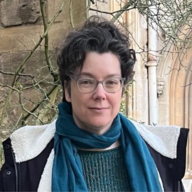 A headshot of Lisa looking at the camera. She has a green jumper on with a blue muffler around her neck. There is a sandstone building in the background, streaked by a few tree branches.