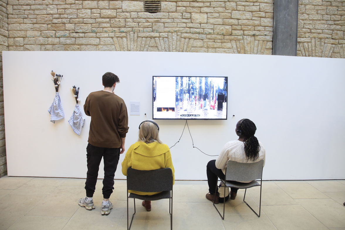3 visitors, one standing and two sitting, watch a film on a video screen