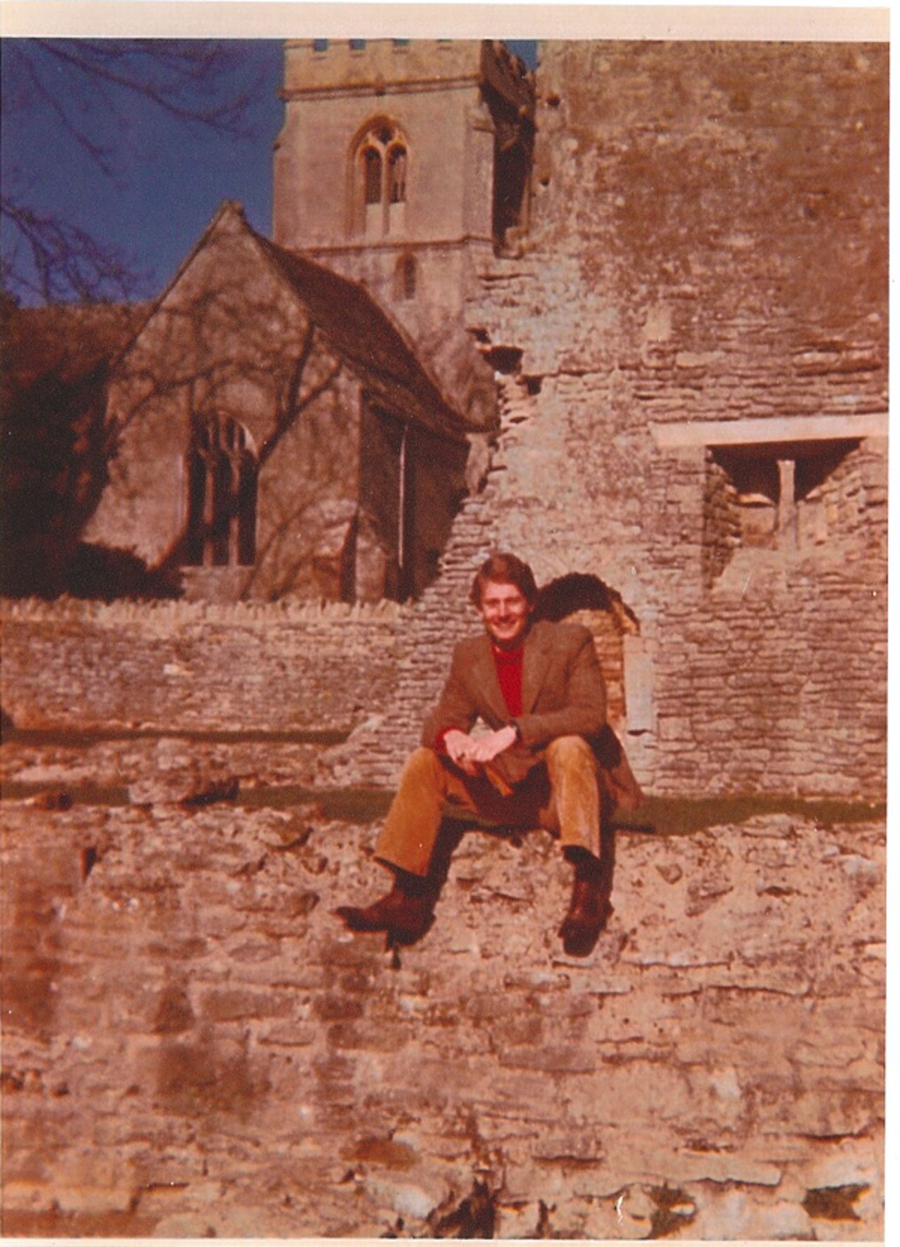 The picture shows Mike sitting on a crumbling stone wall. Behind him is a stone building, possibly that of an old church. He is wearing a warm coat, red sweater and ochre pants.