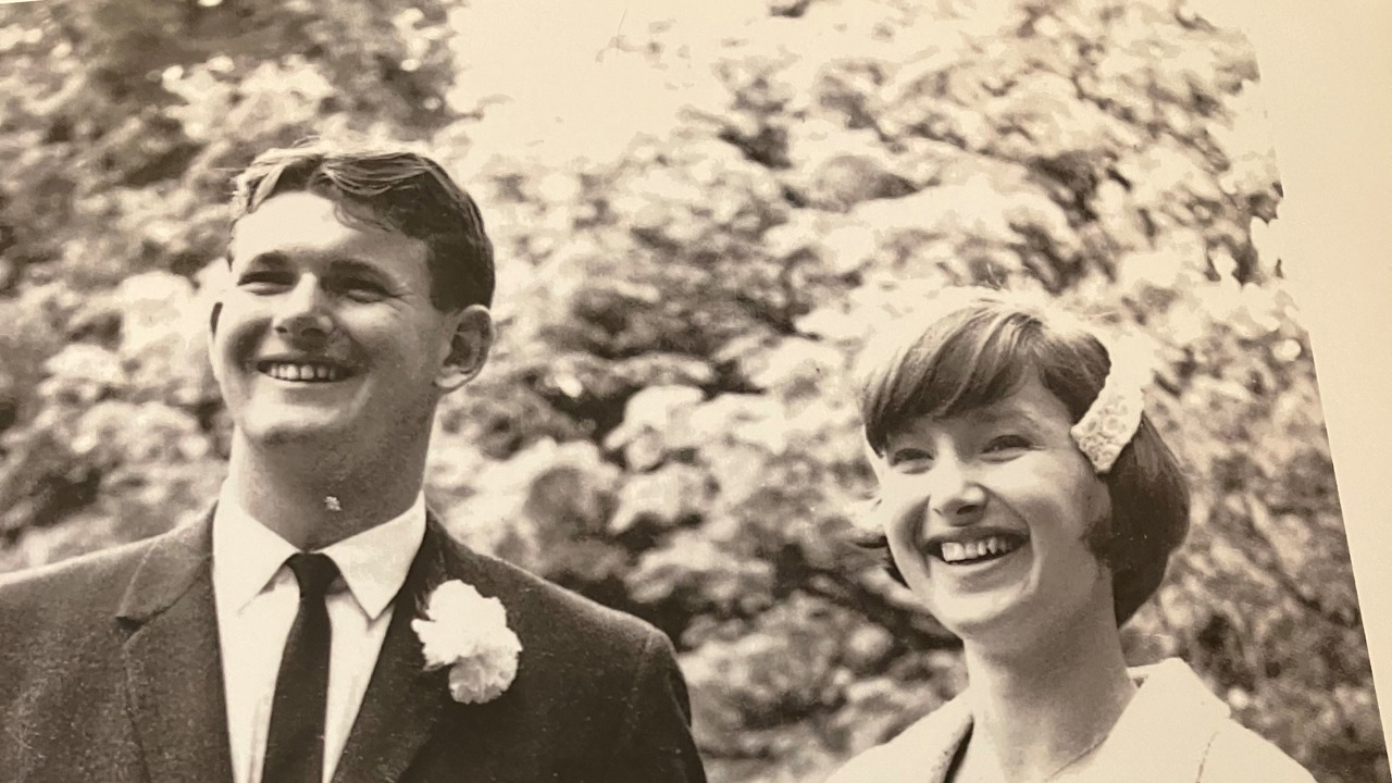 A black-and-white picture of Nick standing with his wife. Nick is in a suit and his wife is wearing a white dress. There are trees in the background.