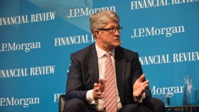 Mike is sat in a suit speaking. Behind him is a blue wall which lists sponsors such as 'J.P. Morgan'