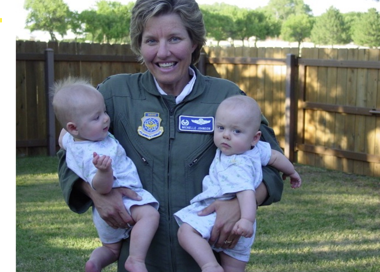 Michelle stood in green uniform in a garden holding her two twin sons (babies) in her arms.