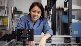 Image of a dark-haired woman in a blue labcoat, smiling at the camera and surrounded by scientific lab equipment.