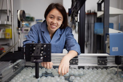 Image of a dark-haired woman in a blue labcoat, smiling at the camera and surrounded by scientific lab equipment.
