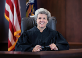 Susan sat at a desk, smiling, wearing a judges robe. American flag behind her. She is holding glasses and wearing large pearl earrings.