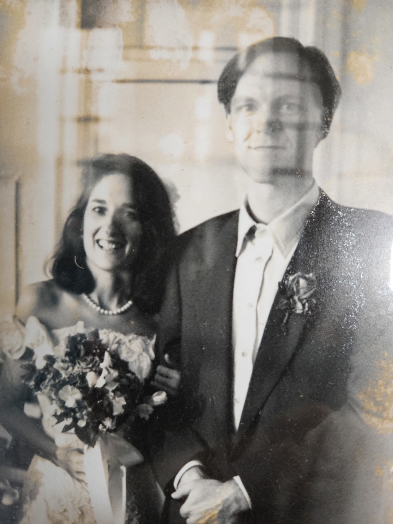 Black and white photo of a married couple. To the left is a lady smiling wearing a white dress, pearl necklace and holding a bouquet. To the right is a young man in a shirt and blazer and rose in the pocket.