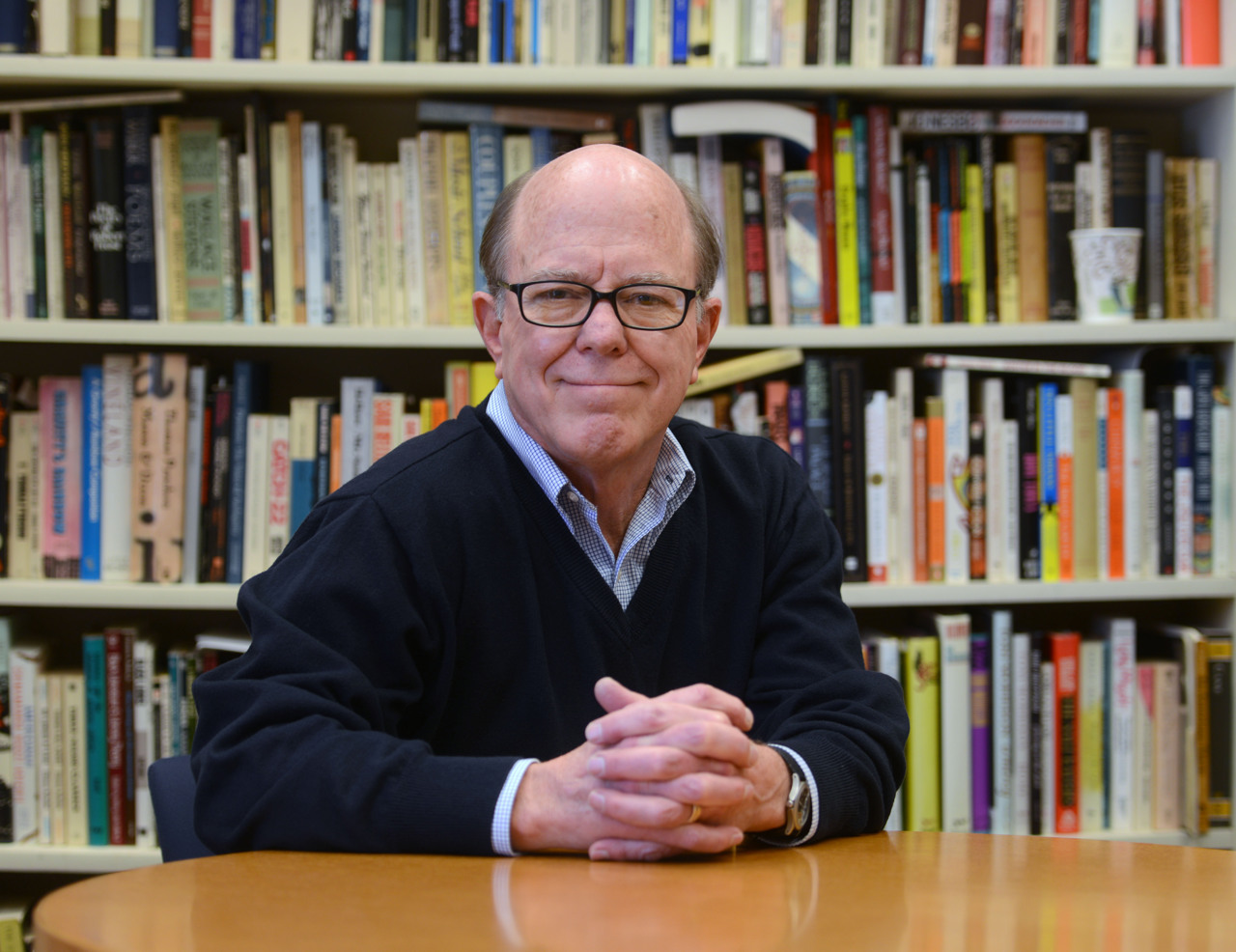 A headshot of George with a bookshelf in the background. George is wearing a dark-blue jumper on a sky-blue shirt,