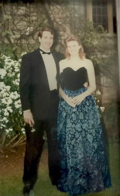 Photo of Randy stood to the left of his wife in front of Rhodes House. Randy is wearing a dinner suit, and Rachel is wearing a long teal and black dress.
